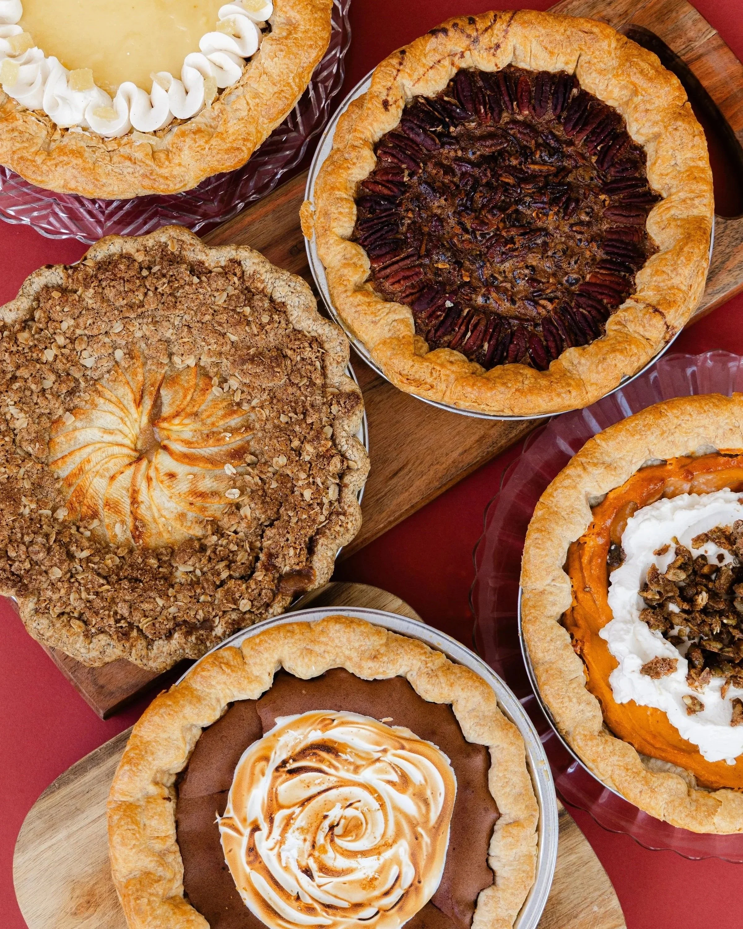 Assorted pies on plates and a wooden cutting board, including a chocolate cream pie, pecan pie, and a crumb-topped pie with a caramel swirl.