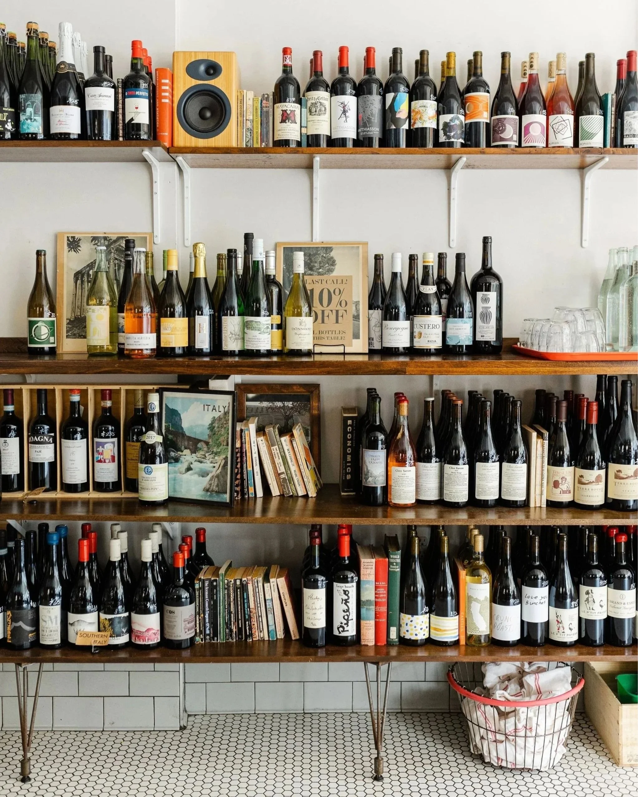 A retail display of various bottles of wine on three wooden shelves against a white wall. The display includes wine bottles, books, a speaker, framed pictures, and a sign offering a 10% discount. The lower shelf has a wire basket with a cloth inside and is located in a vintage-style tiled room.