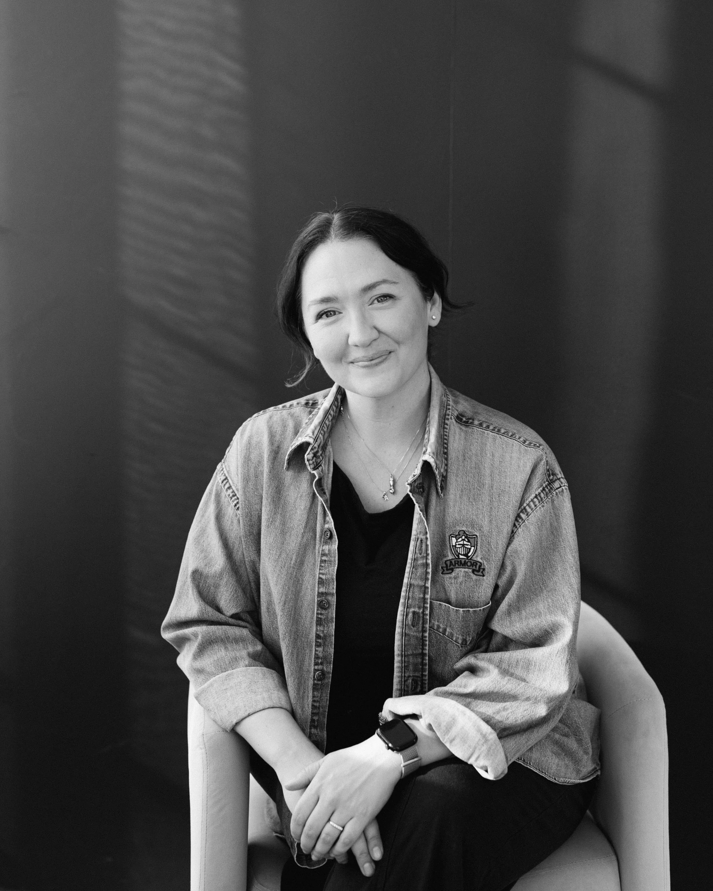 Black and white photo of a smiling woman with short dark hair, wearing a denim jacket with a badge on the pocket, a black shirt, an Apple Watch, and a ring.