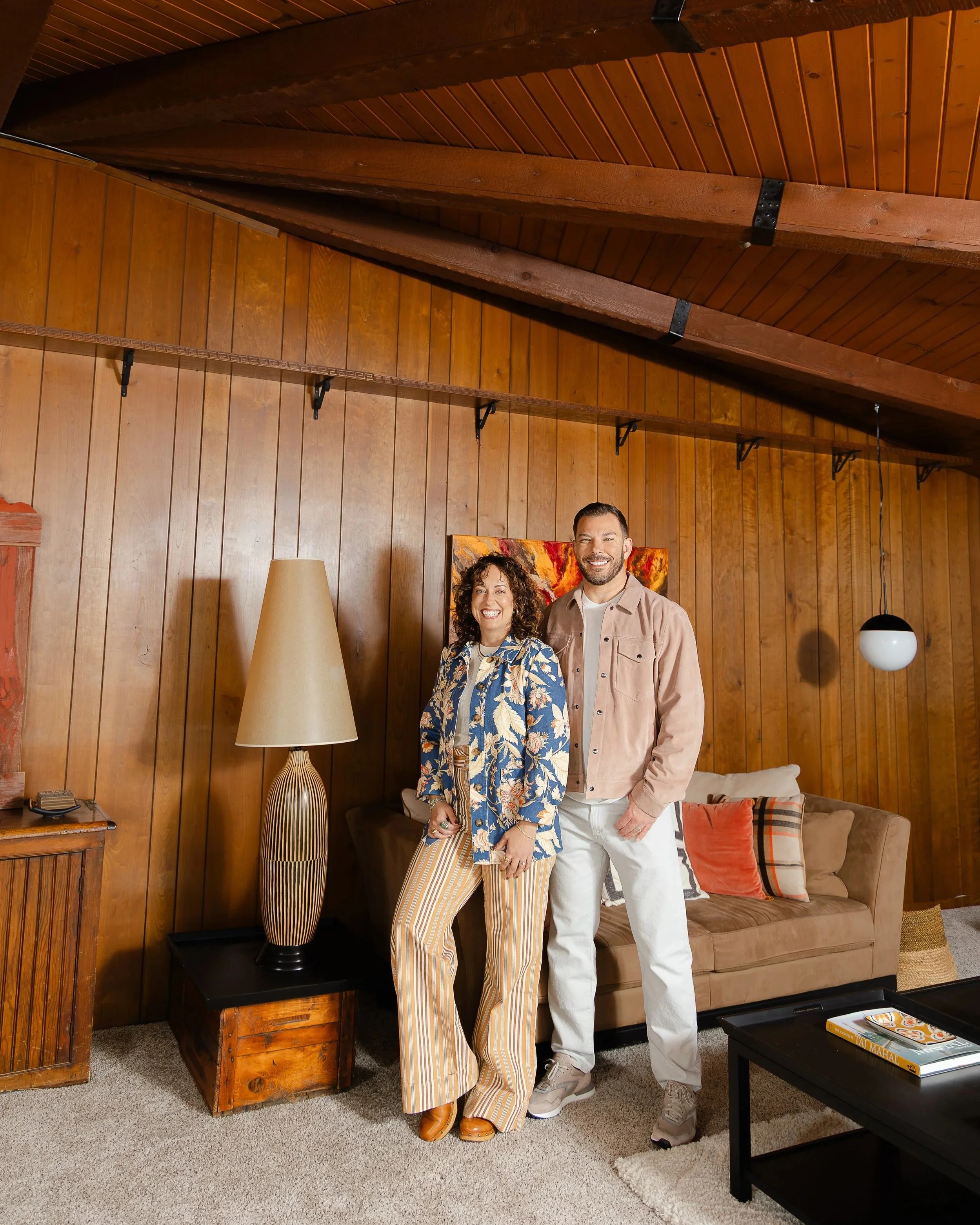 Two people, a woman and a man, standing in a living room with wood-paneled walls, posing for a photo while smiling. The woman is wearing striped pants and a floral blouse, and the man is in light pants and a beige jacket. Behind them, there is a brown couch with colorful pillows, a painting on the wall, and a wooden table with books and a tray on it.