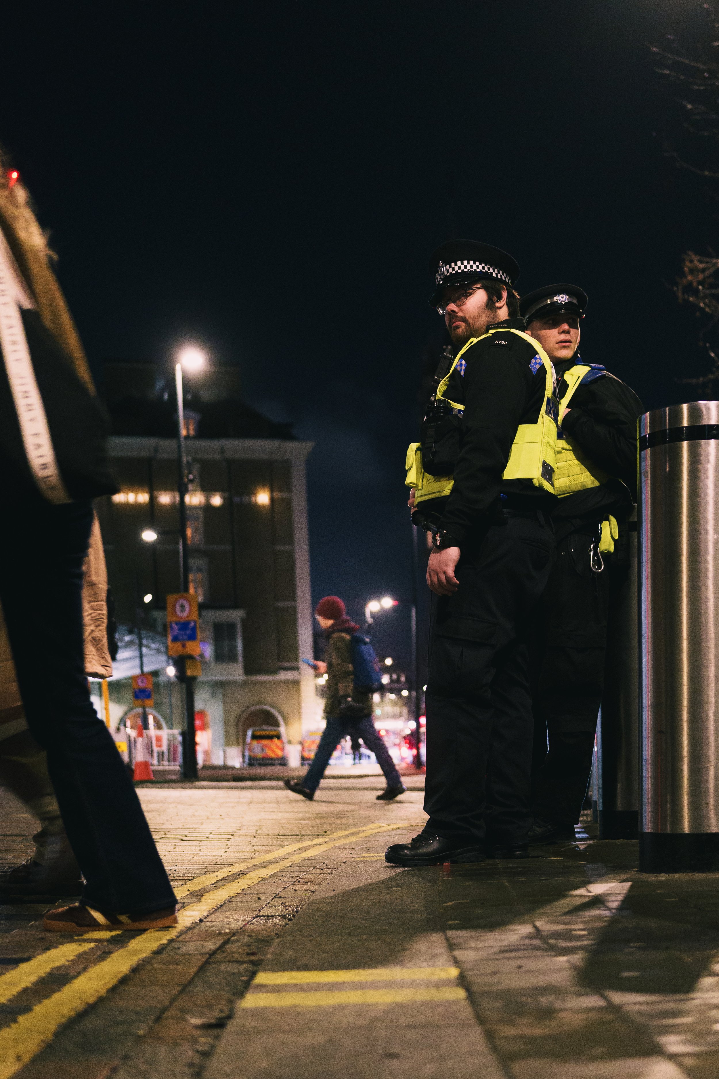 Two police officers in reflective vests and hats standing on a city sidewalk at night, with pedestrians walking by and buildings lit in the background.