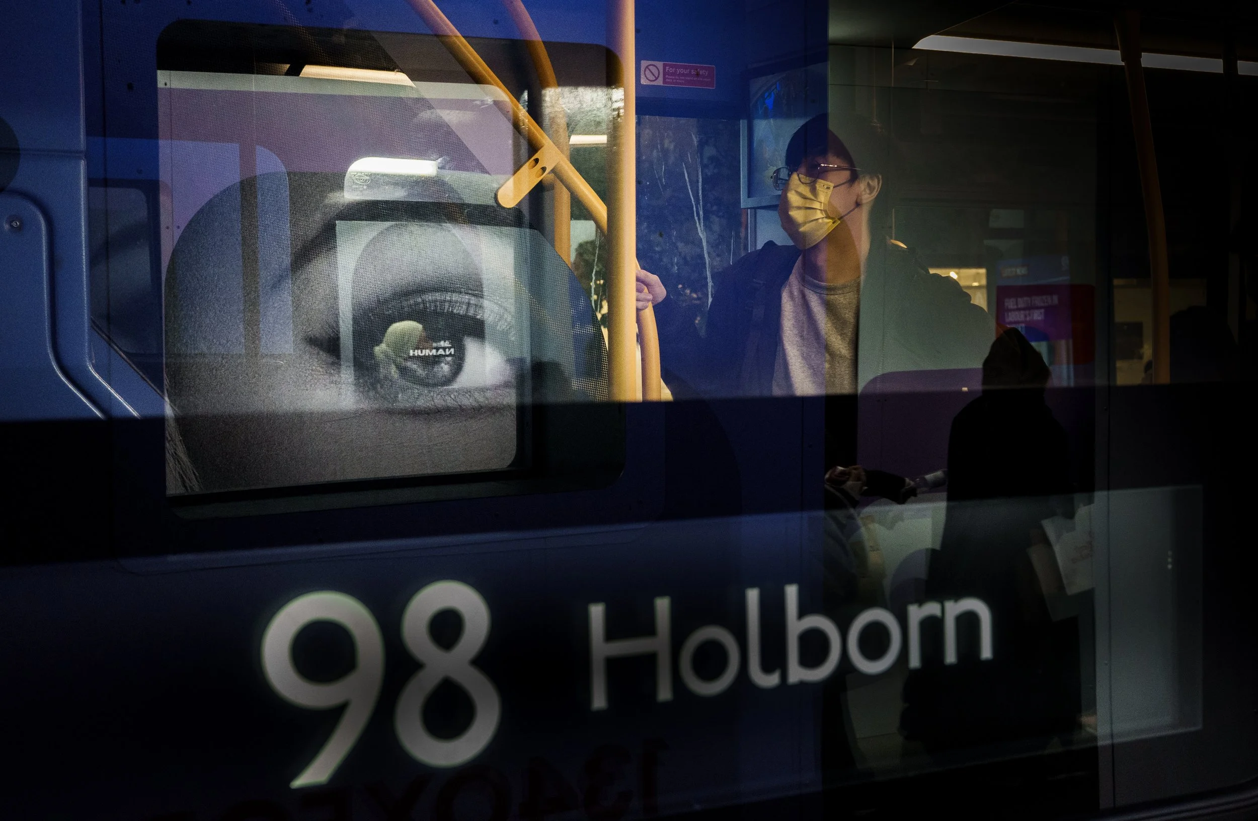 A person wearing a yellow face mask and gray shirt standing inside a bus, holding a yellow handrail, with the reflected image of a large eye on the bus window, and a sign indicating the bus route 98 to Holborn.
