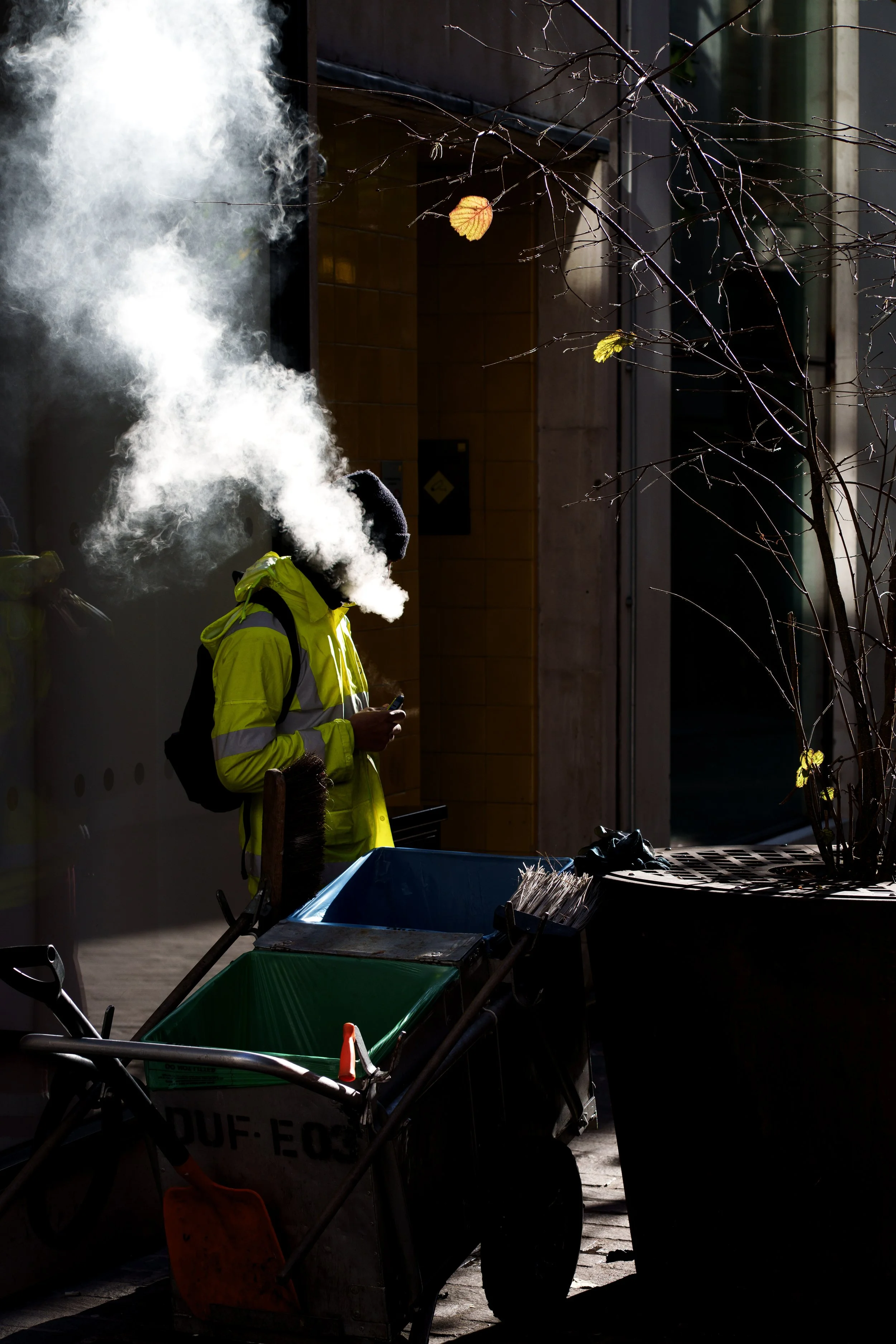A person wearing a yellow reflective jacket and black hat, smoking or exhaling smoke, standing beside trash bins and cleaning tools on a city sidewalk during the daytime.