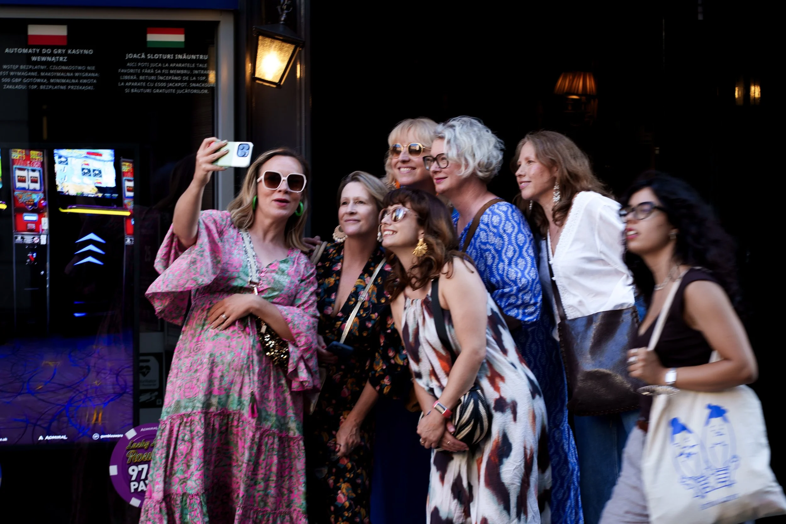 A group of women and one girl taking a selfie together outside a building. They are dressed in colorful clothing and appear to be happy.