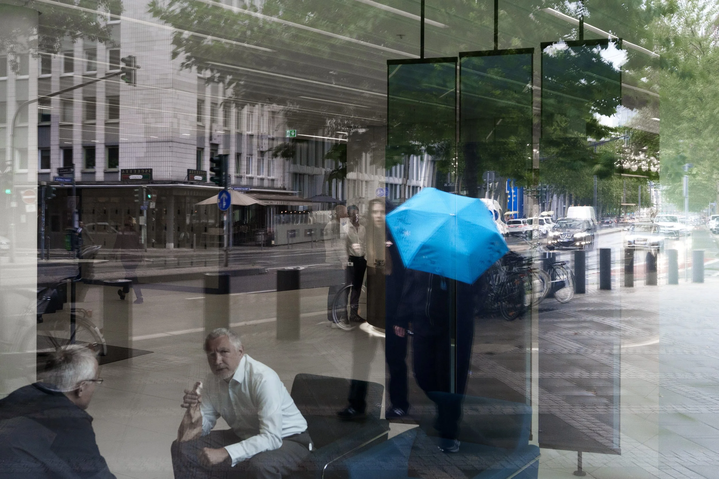 Reflection of people and city street scene seen through glass window, showing individuals walking on sidewalk, parked bikes, and commercial buildings across the street, with some people sitting inside.