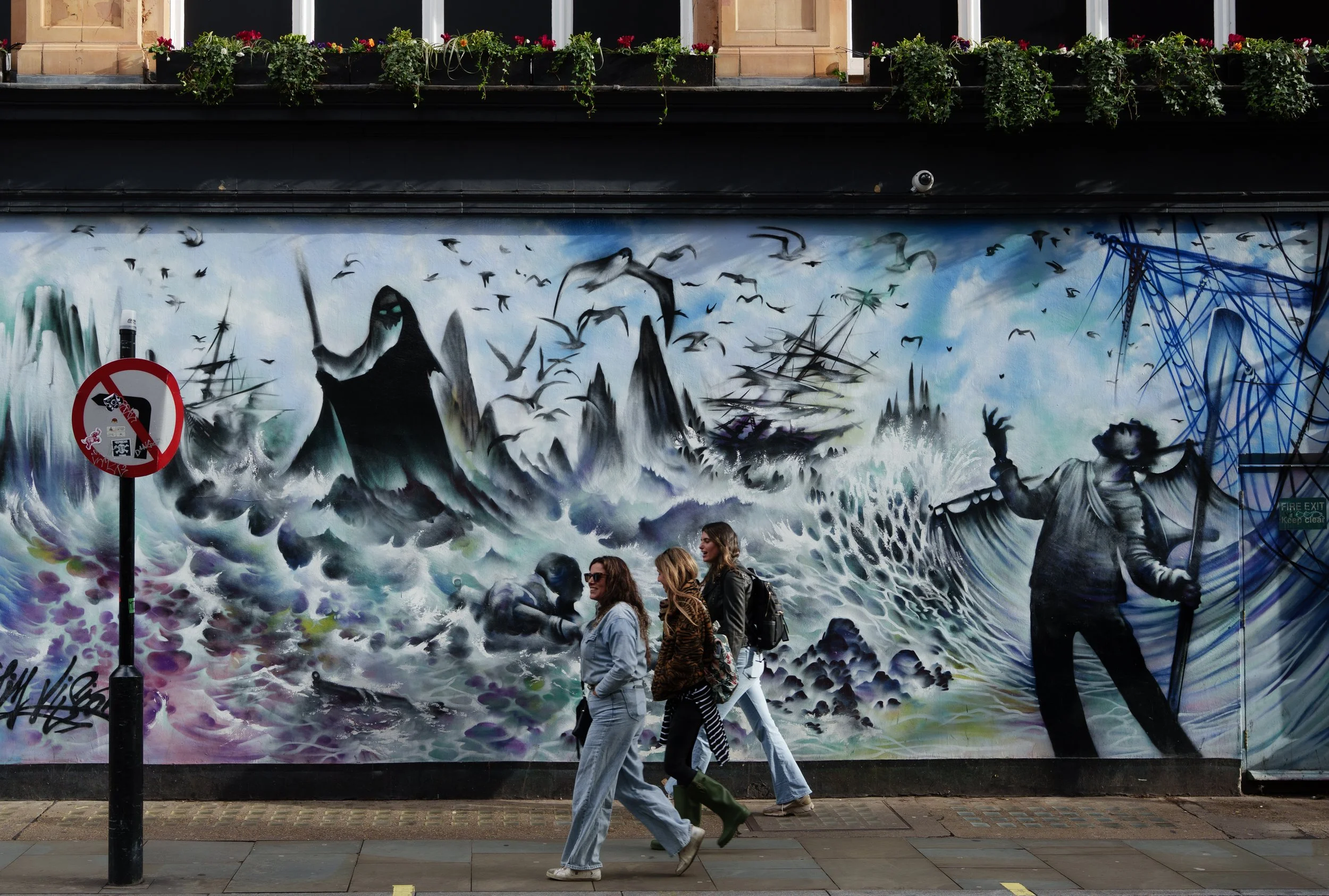 Street art mural depicting a stormy sea with waves and dark mountains, a large figure with a staff, and birds flying in the sky, with three women walking by in the foreground.