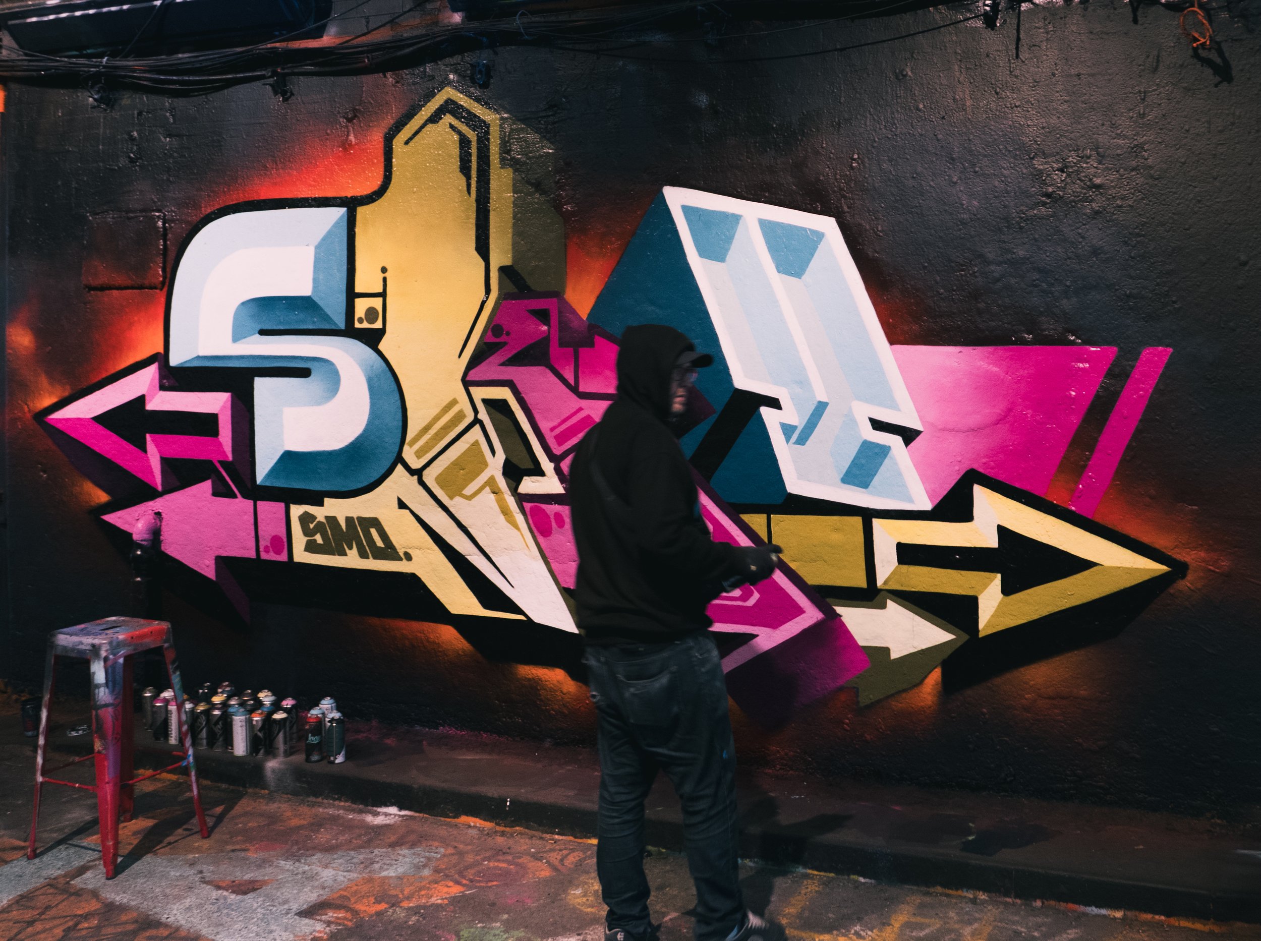 A man in a black hoodie and jeans standing in front of a colorful graffiti mural on a black wall, with spray paint cans on the floor to the left.