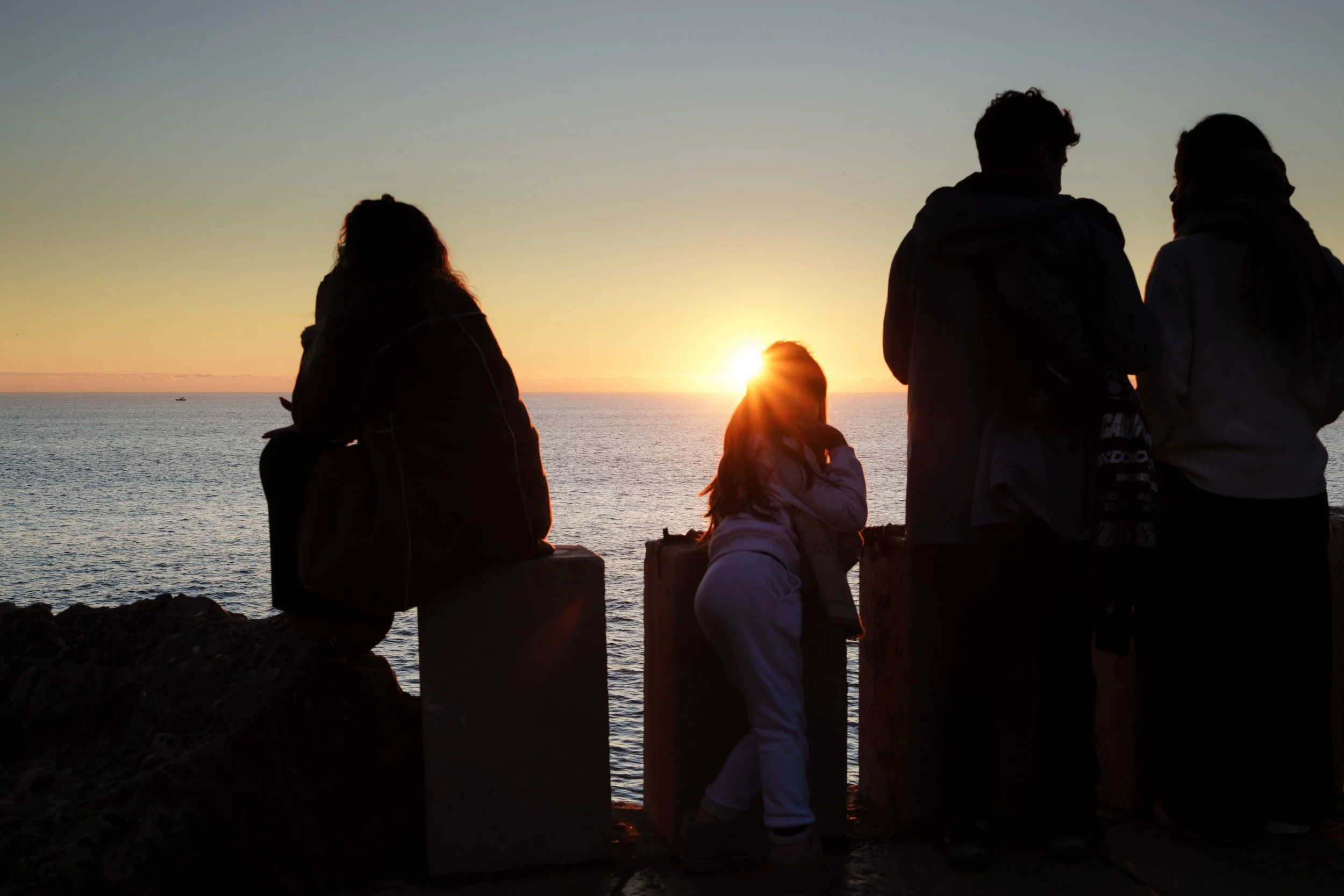 Silhouettes of five people, including children, watching a sunset over the ocean from a rocky vantage point.
