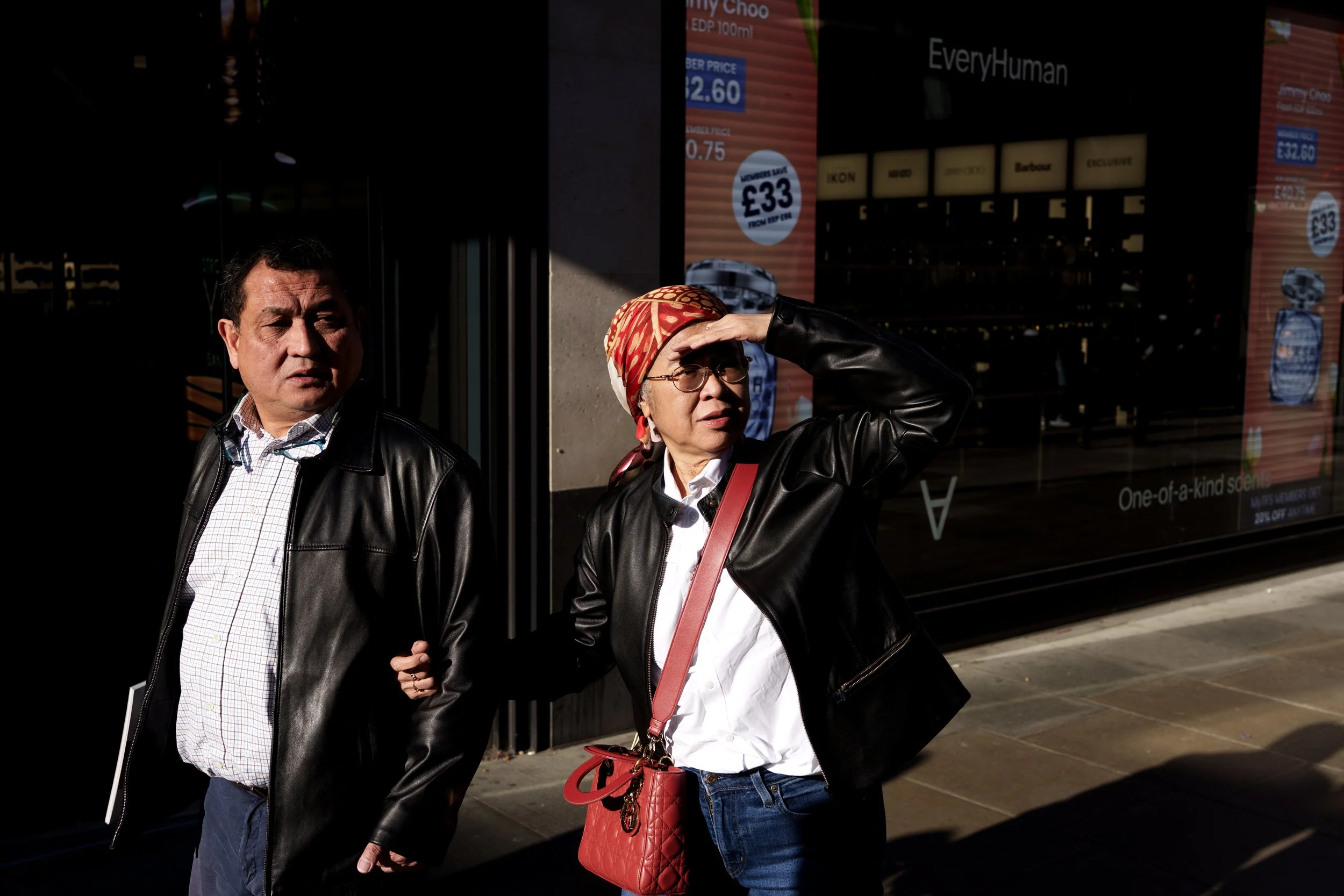 Two people, a man and a woman, standing on a city sidewalk in front of an advertisement window, with the woman shielding her eyes from the sun.