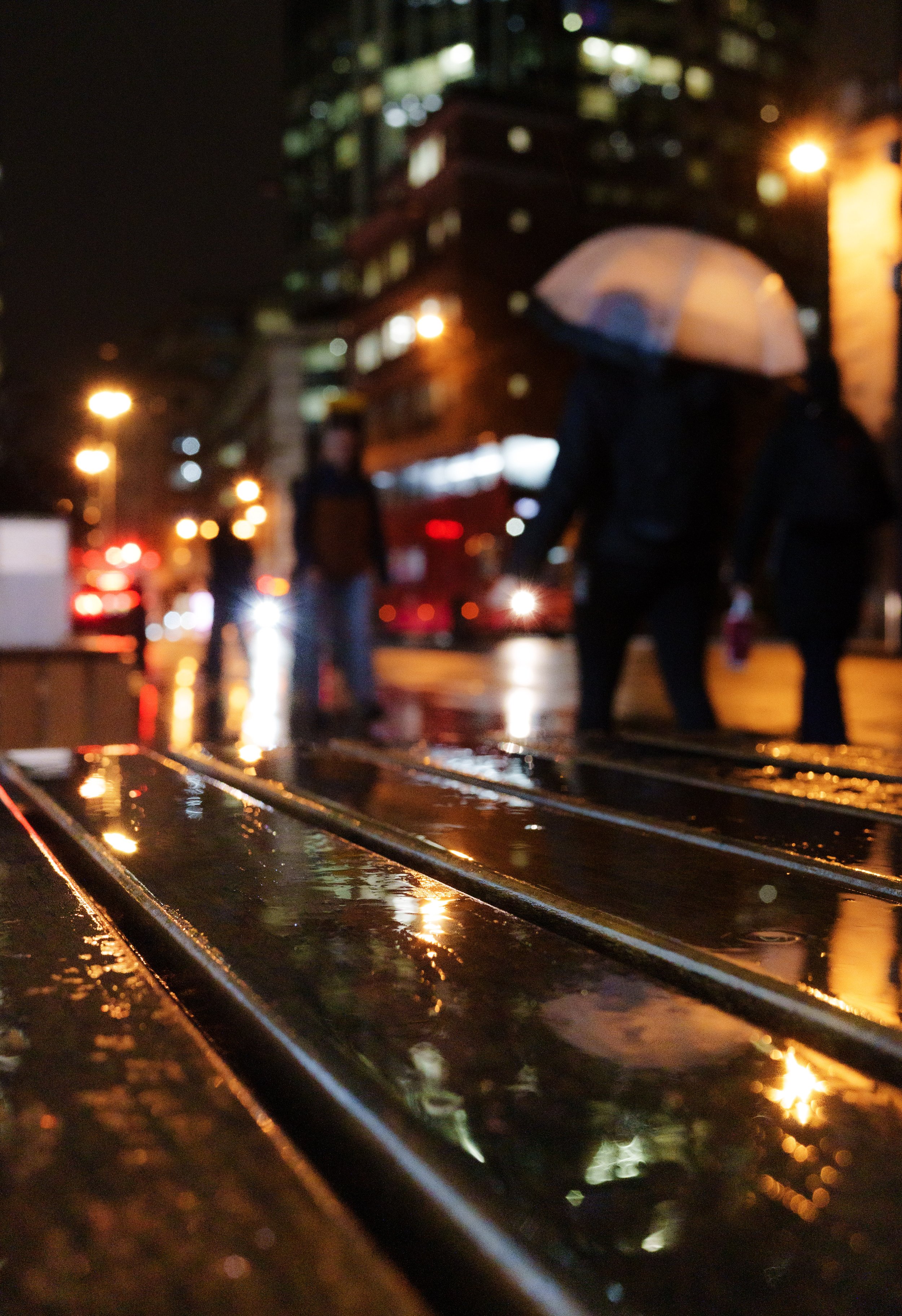 Night scene of people walking on a wet city street with reflections of city lights on the surface, some carrying umbrellas, with blurred buildings and streetlights in the background.