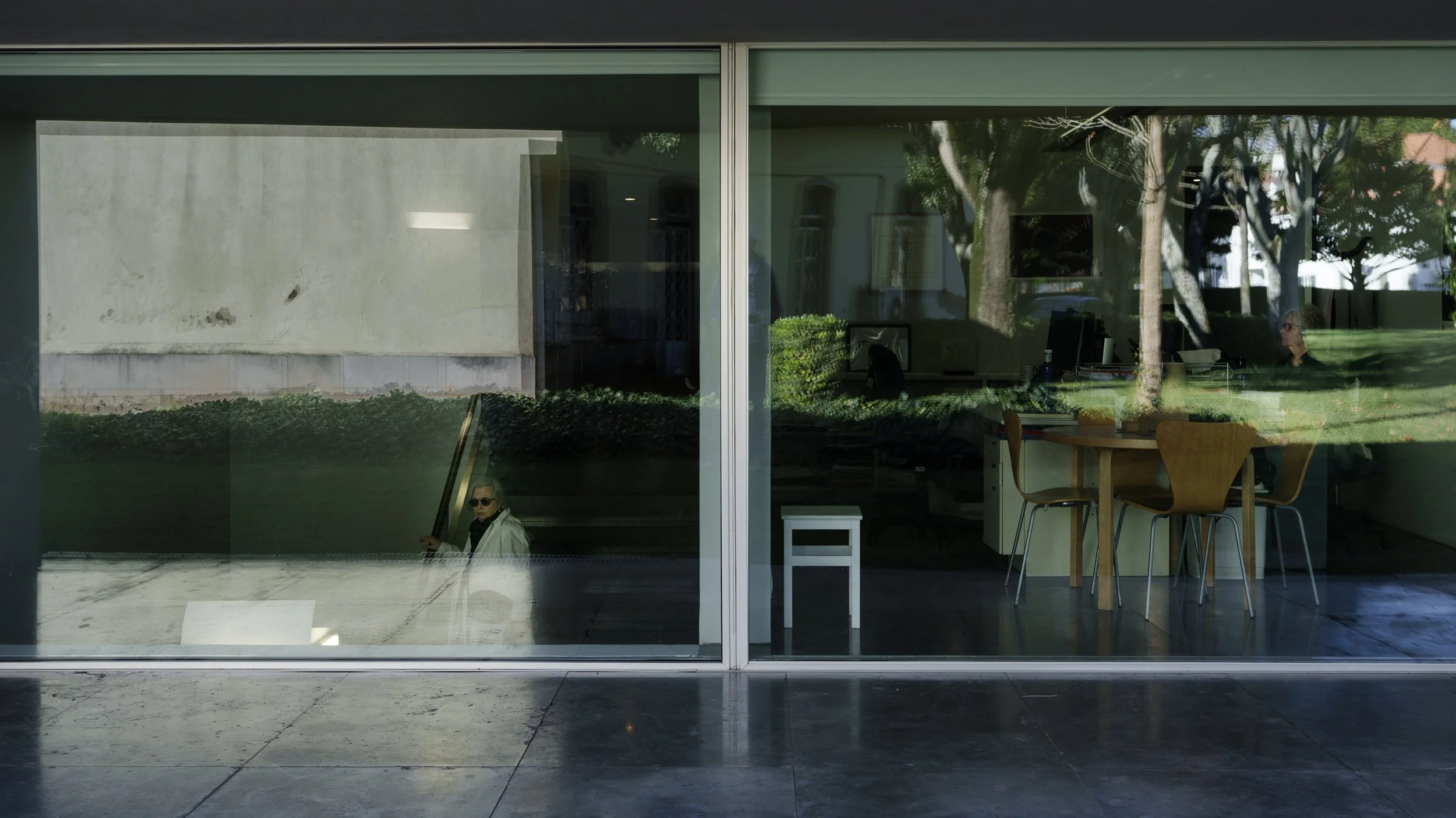 View through a large glass window showing a person sitting inside the building and the reflected outdoor scene with trees, grass, and a woman walking outside.