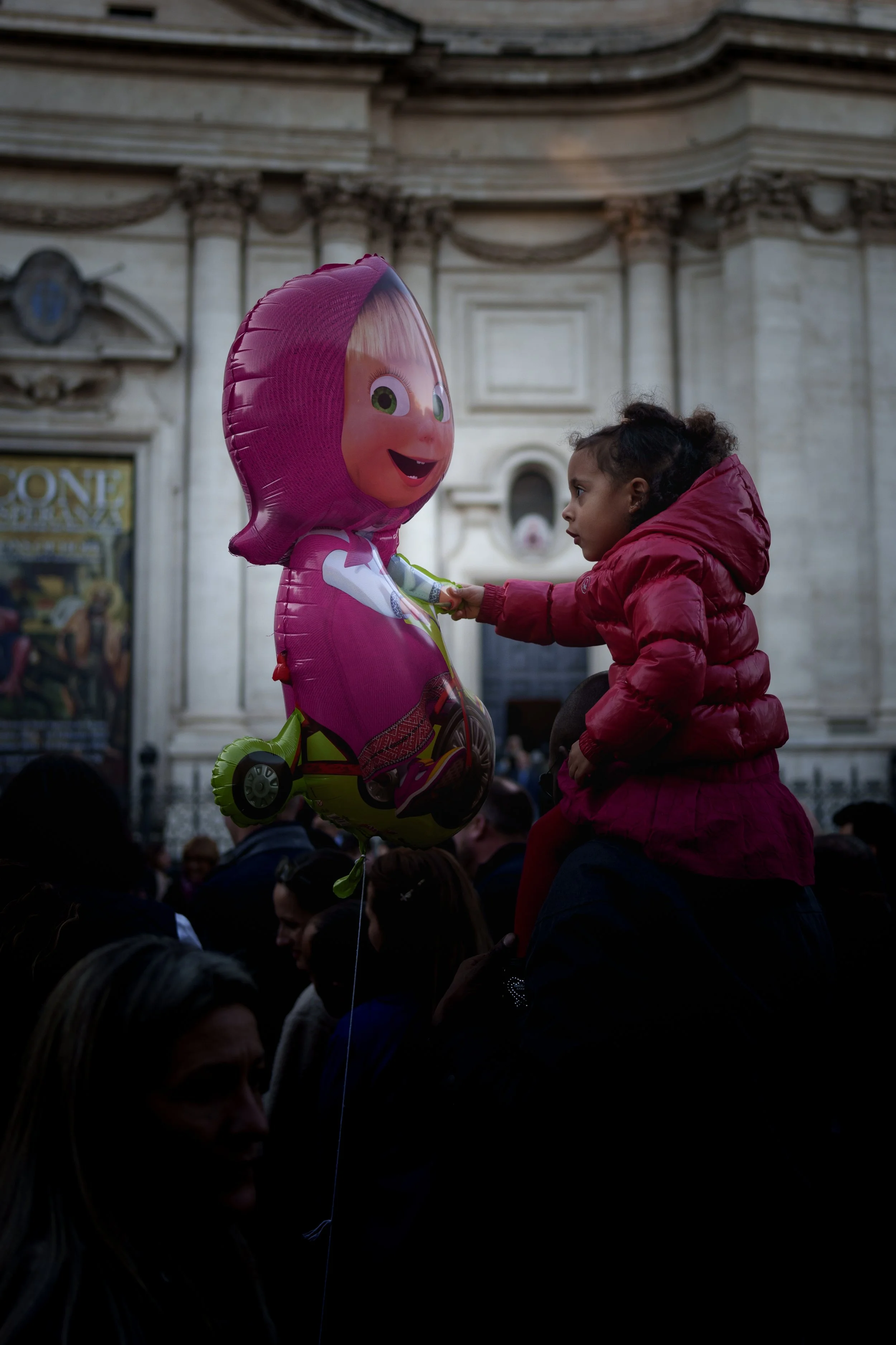 A young girl in a pink jacket is sitting on someone's shoulders, reaching out to touch a pink character balloon with a smiling face, in front of a historic building with ornate architecture.
