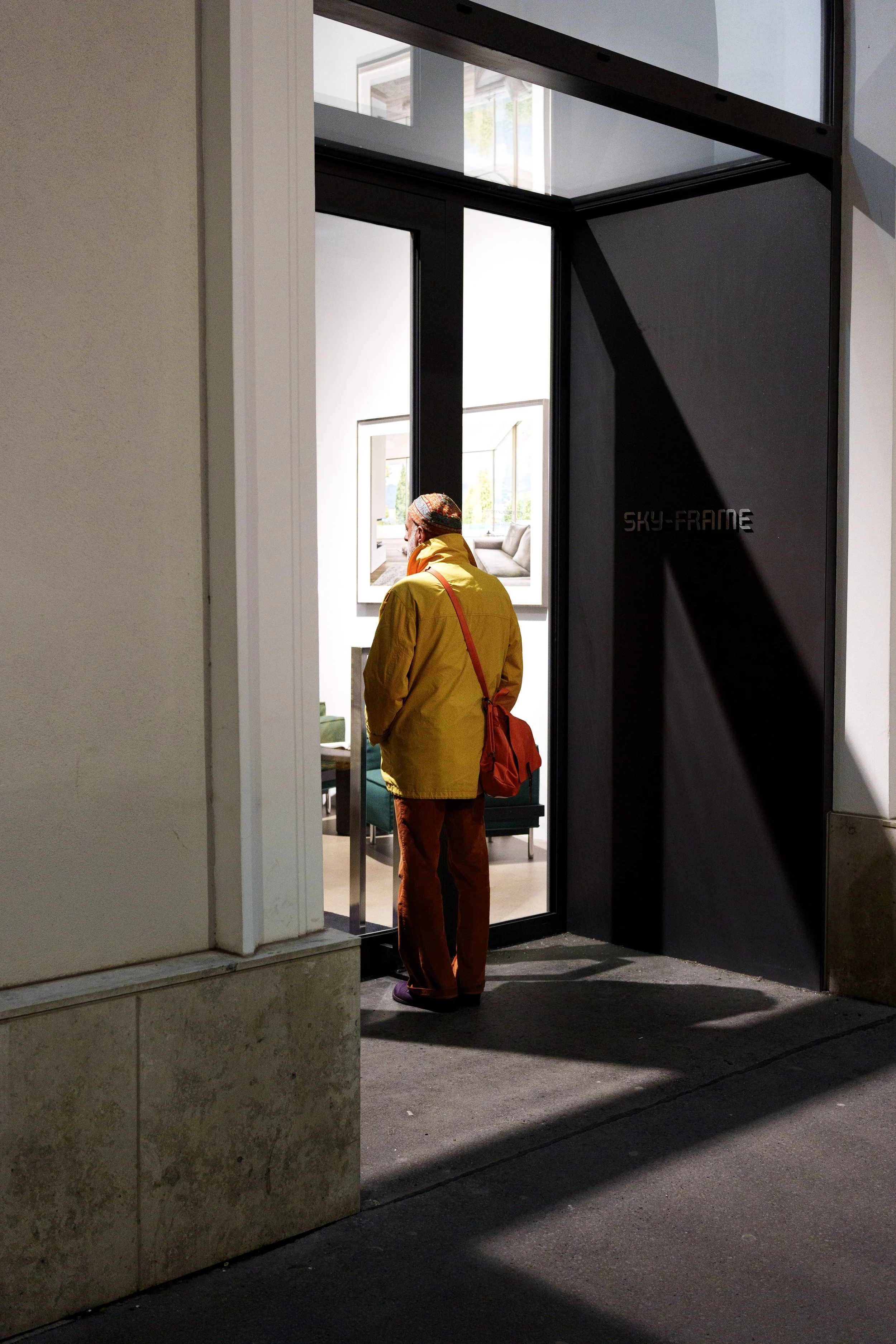 A person in a yellow jacket and red pants stands at the entrance of a building looking inside, shadow cast on the sidewalk, with art or framing visible inside.