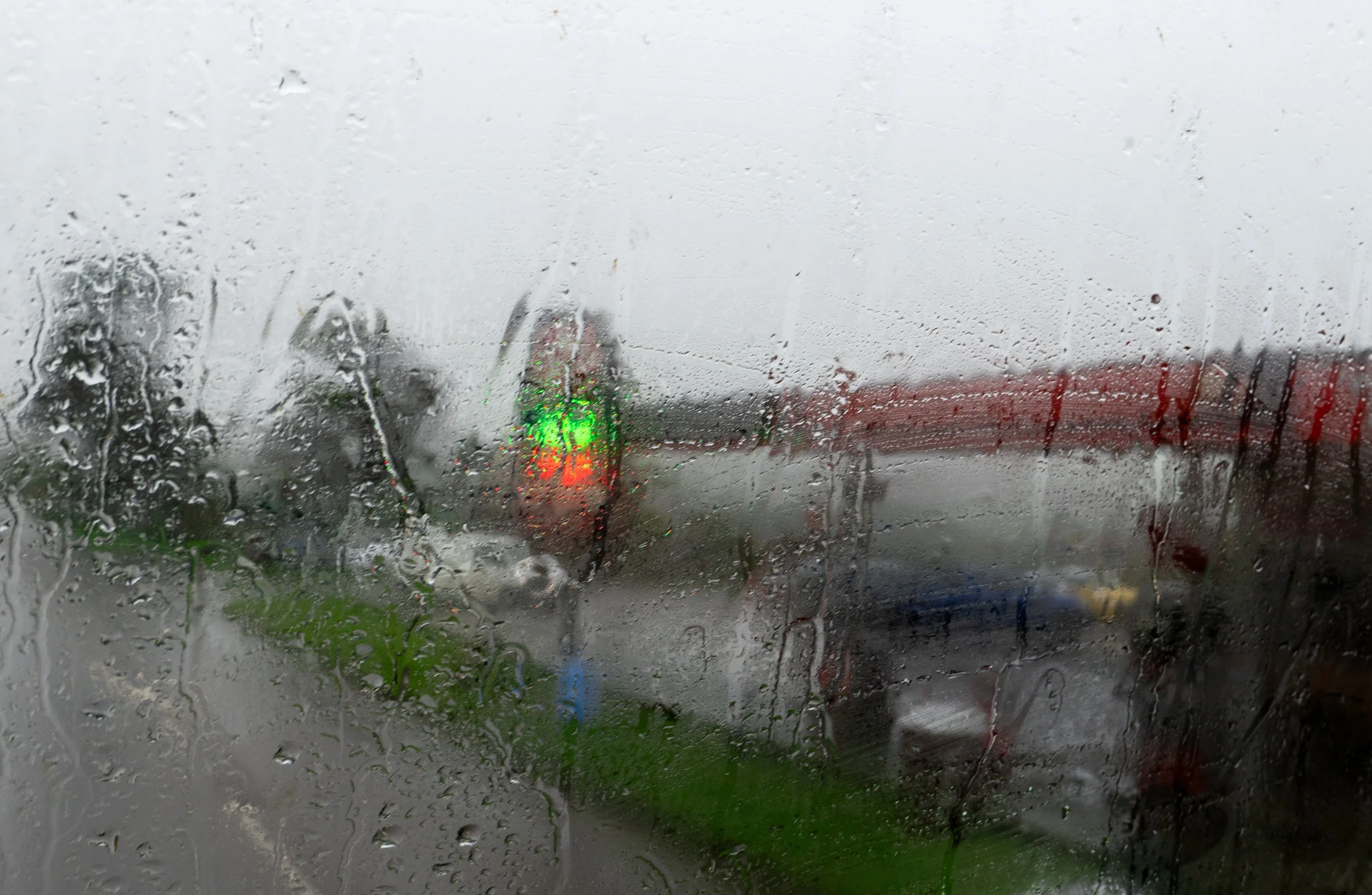 Rainy view through a rain-covered window showing wet roads, blurry trees, and traffic signals, with a red bridge in the background.