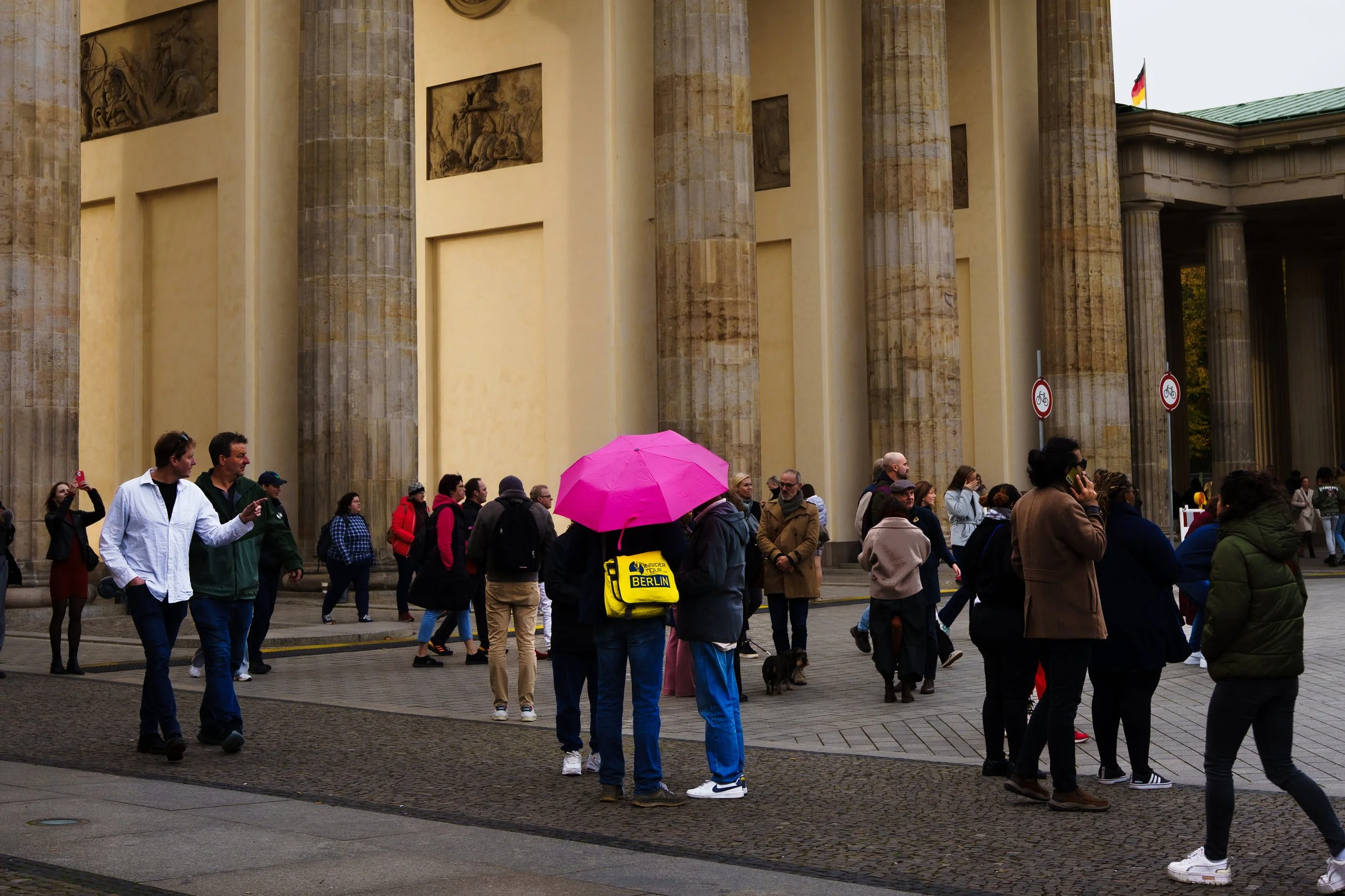 People gathered outside a historic building with large columns, some talking on phones, others walking or standing, one person with a bright pink umbrella, others with backpacks and jackets, a small dog visible among the crowd, and German flags flyin
