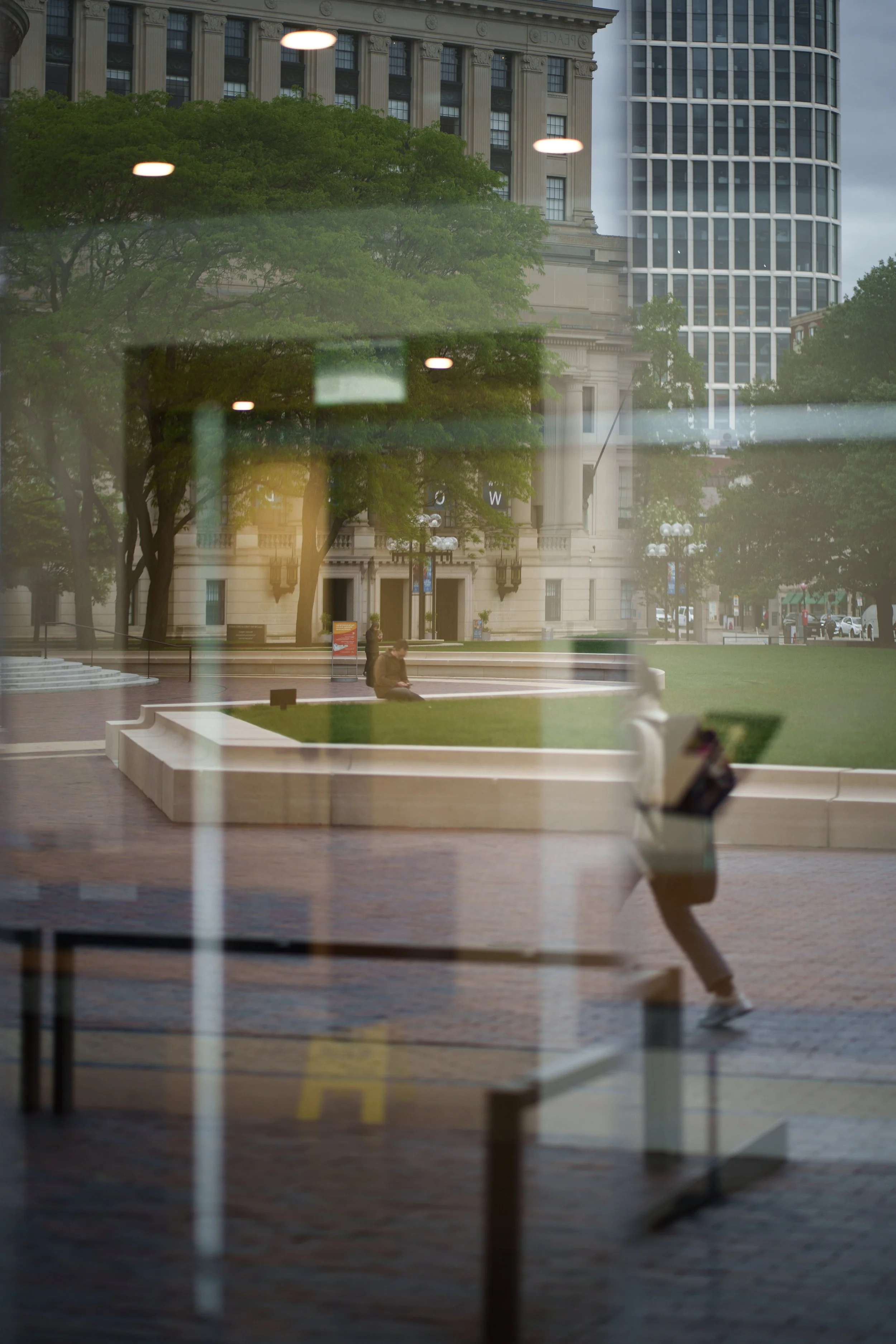 Reflected city park scene with trees, a person sitting on a bench, a person running, and buildings in the background, seen through a glass window.