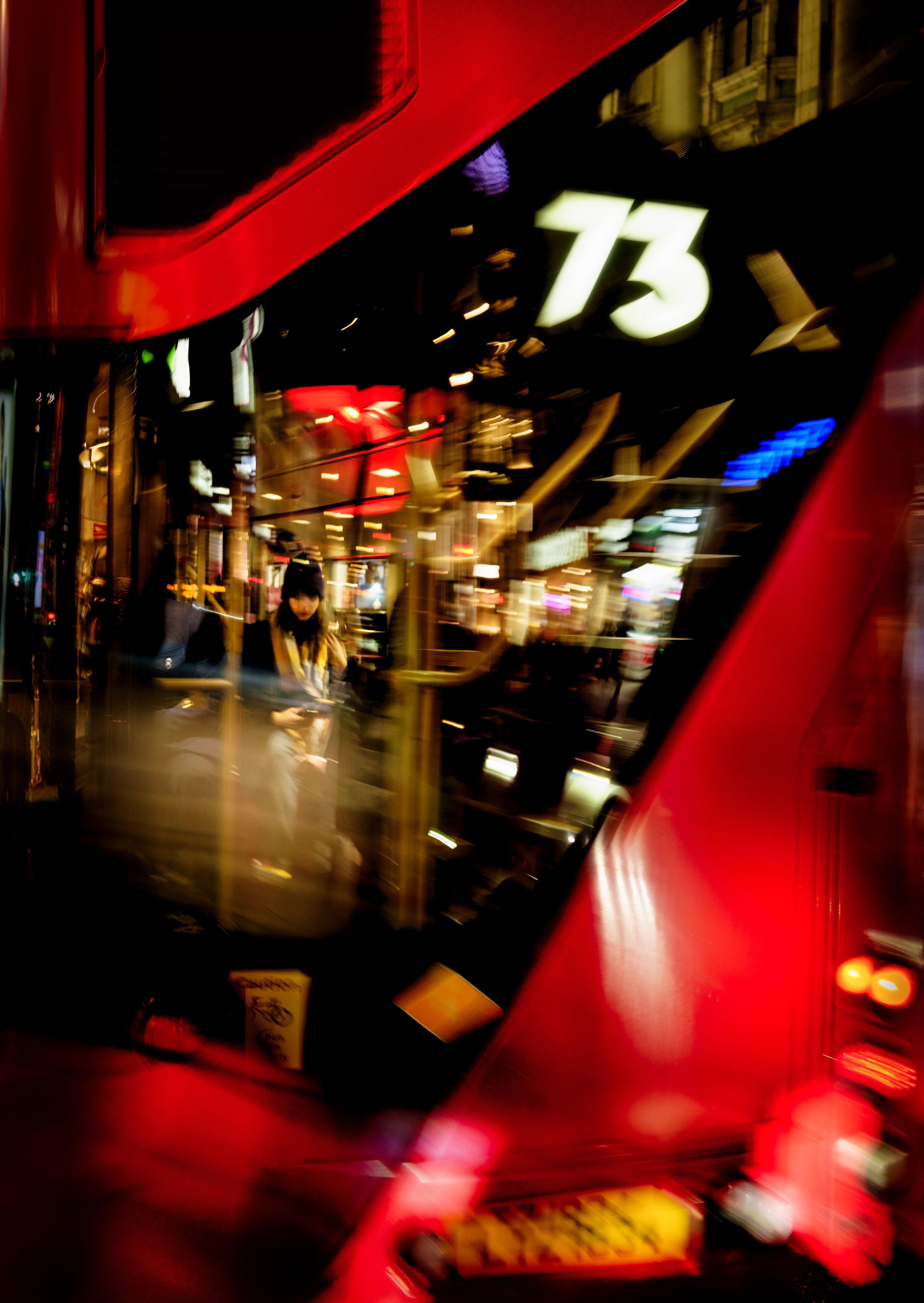 Blurred city street scene at night viewed through the angled front window of a red bus, with city lights and a woman seen sitting inside the bus.