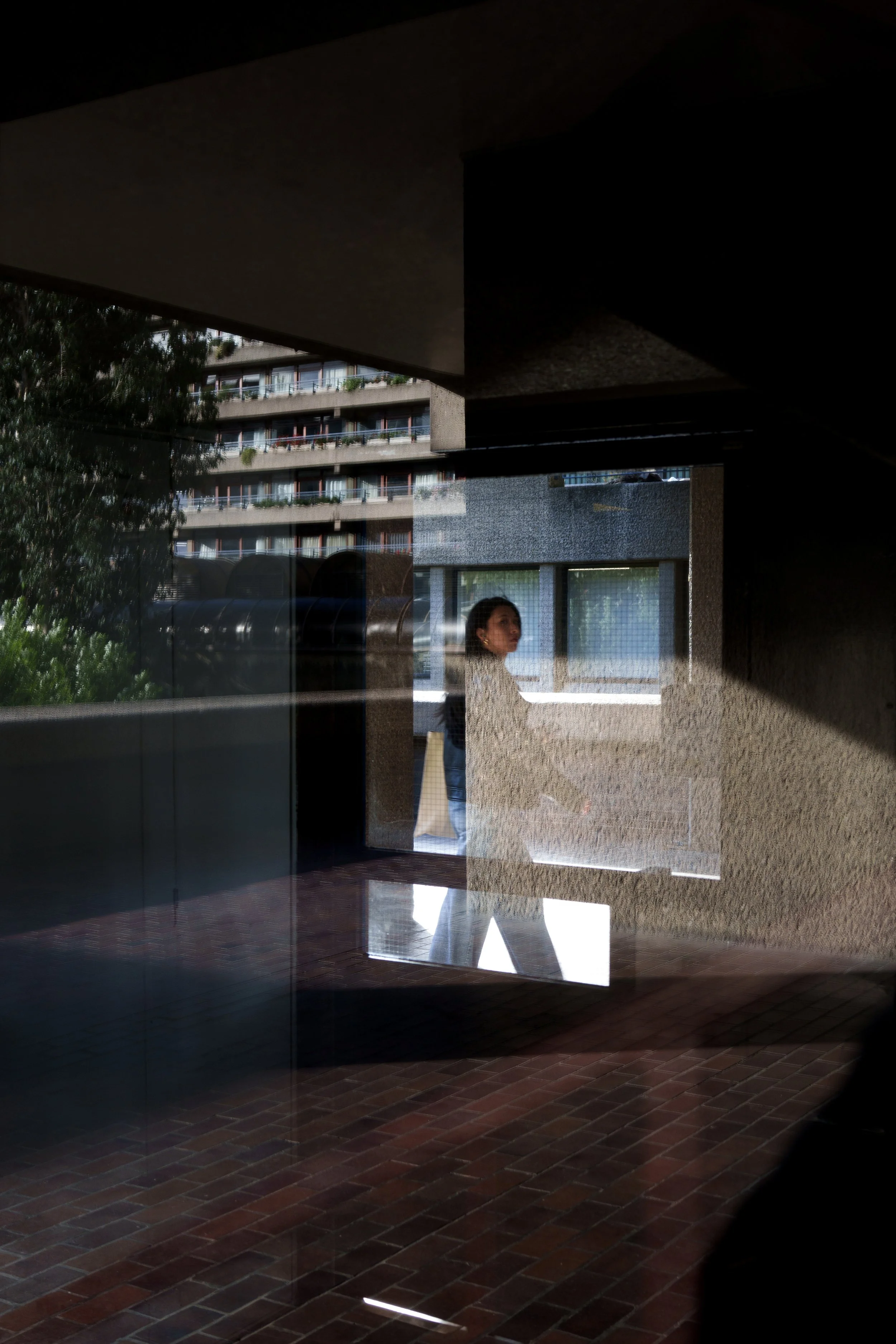 A woman walking past glass reflection on an apartment building's glass window, with trees and a brick pathway visible outside.