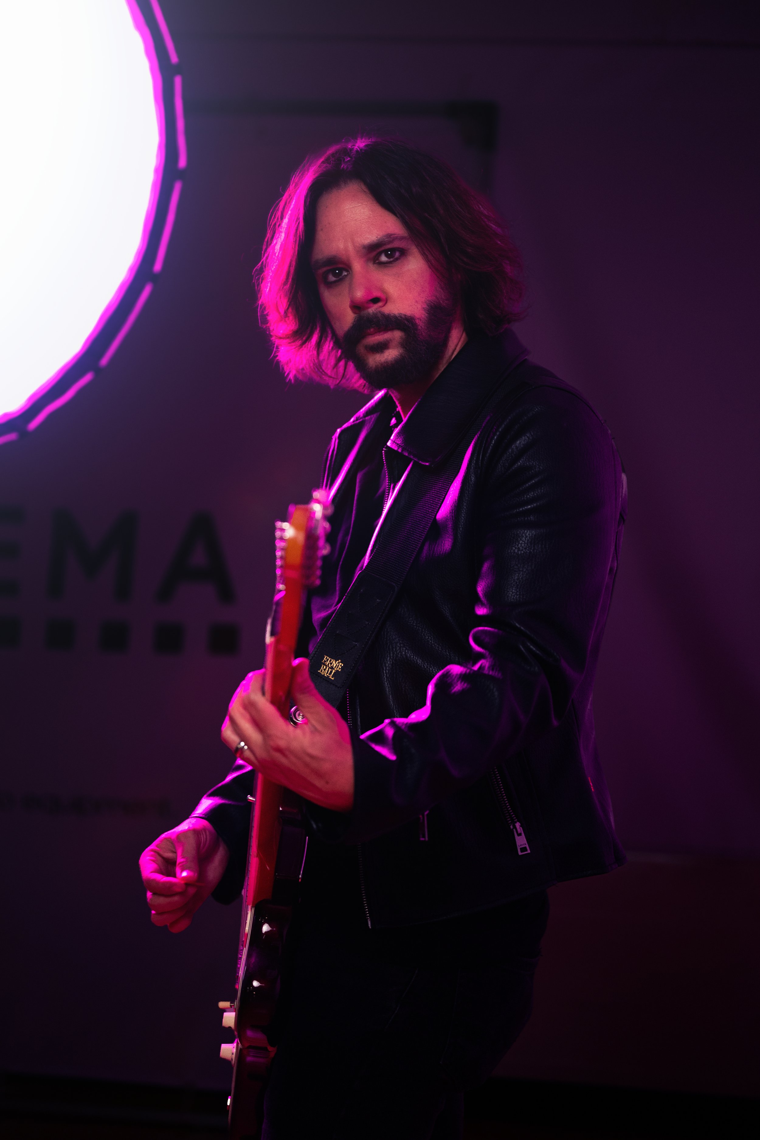 Man with long dark hair and beard, wearing a black leather jacket, holds a guitar, standing against a dark background with purple neon lighting.