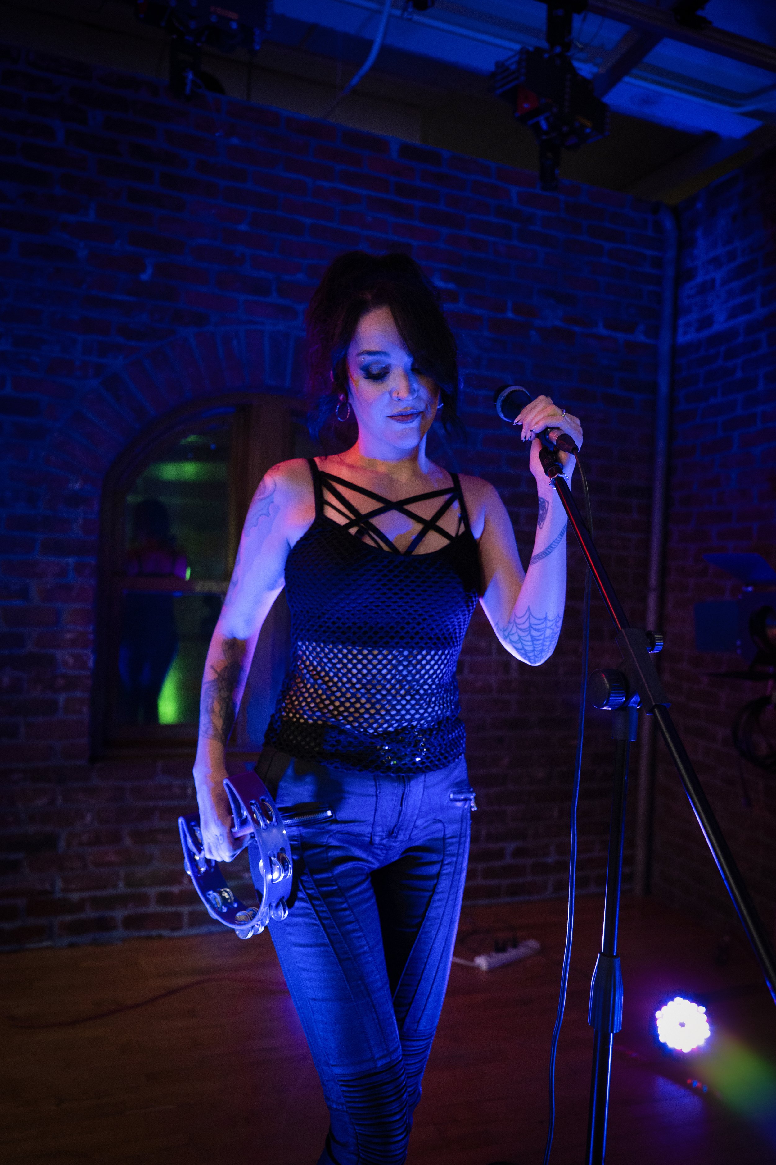 A woman performing on stage with a tambourine in one hand and a microphone in the other, in a dimly lit setting with blue and purple lighting, exposed brick wall background.