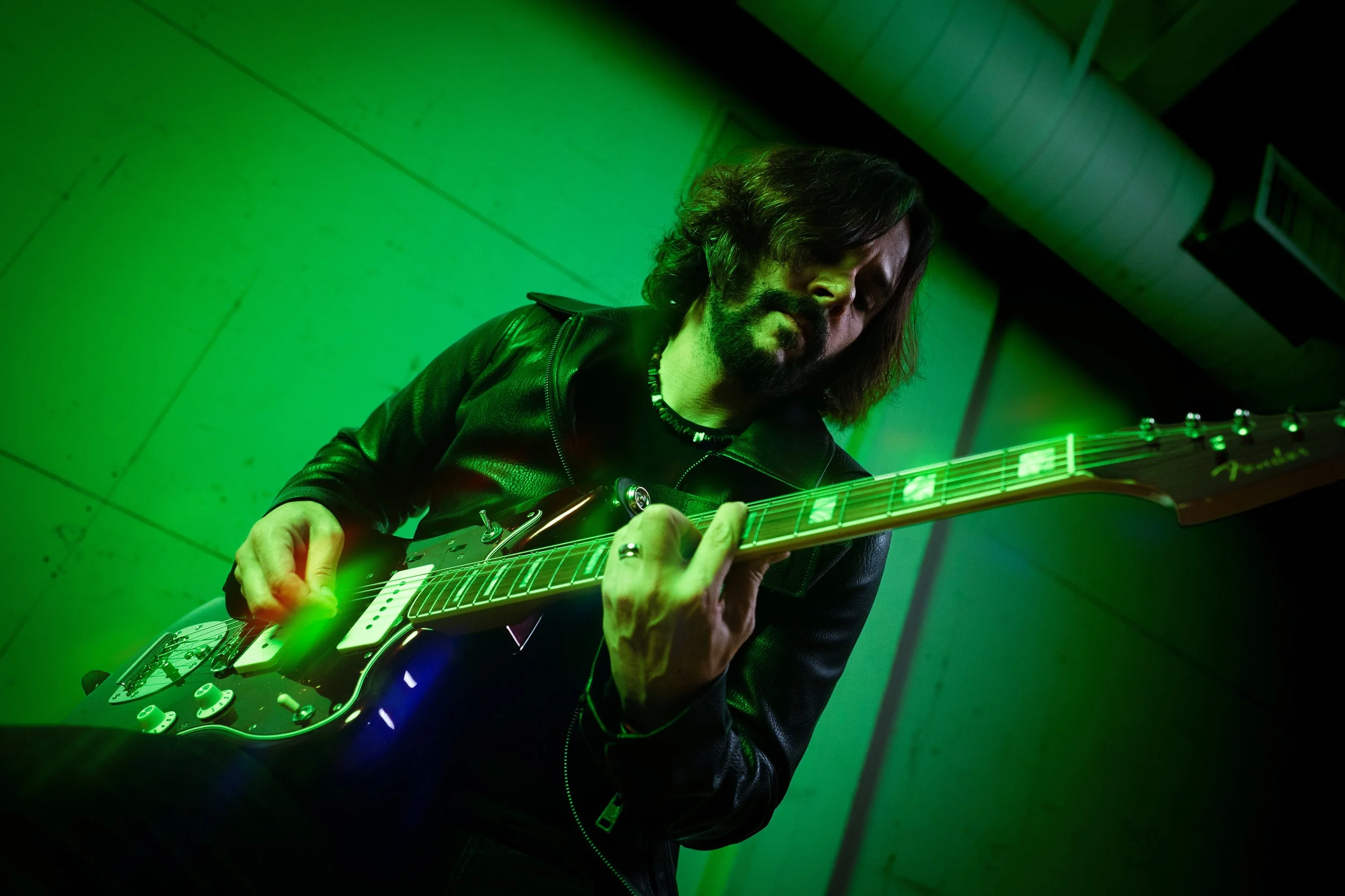 A person with long hair and a beard playing an electric guitar in a room with green lighting.