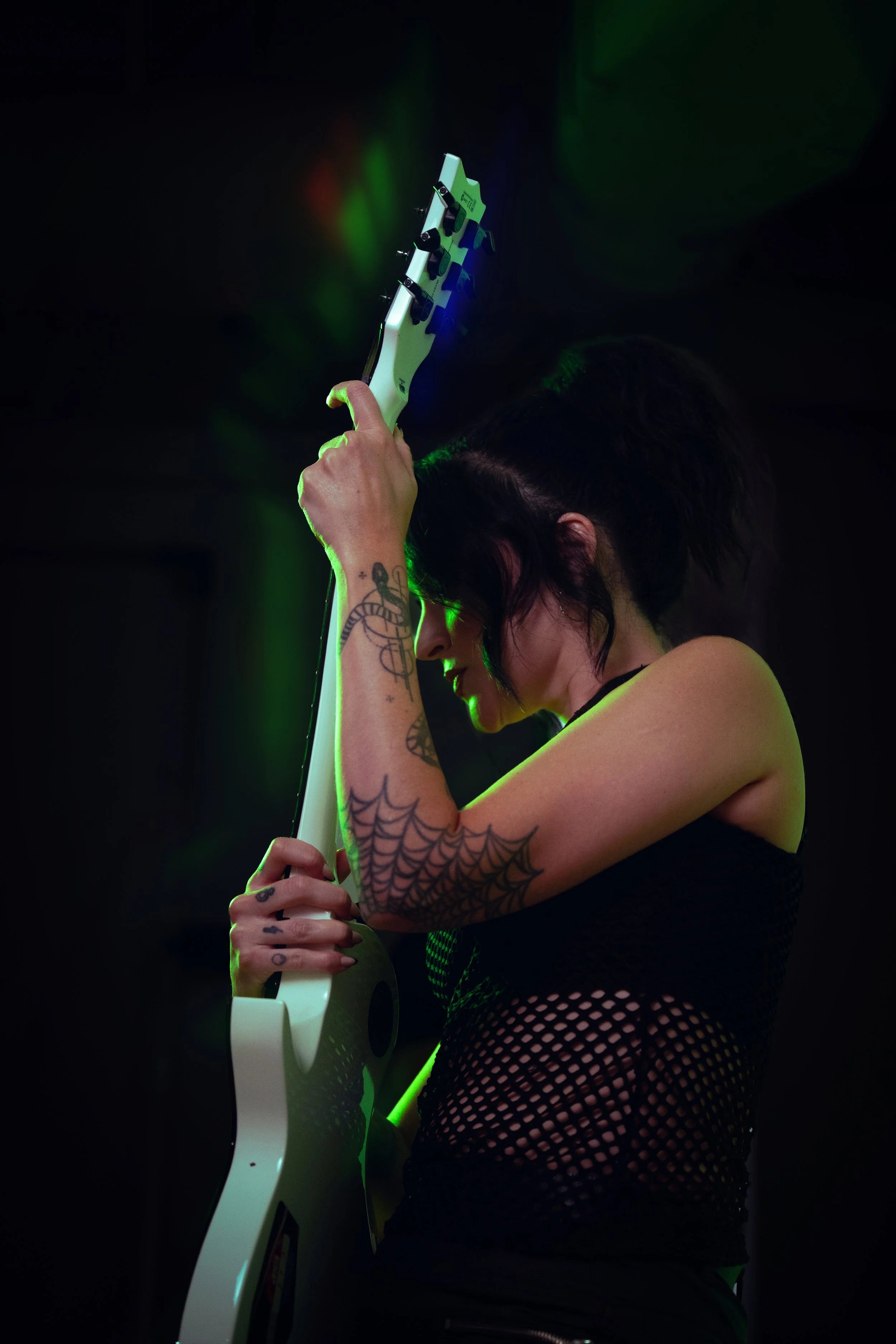 A woman with black hair and tattoos on her arms holding a white electric guitar in a dark room with green and red lights.