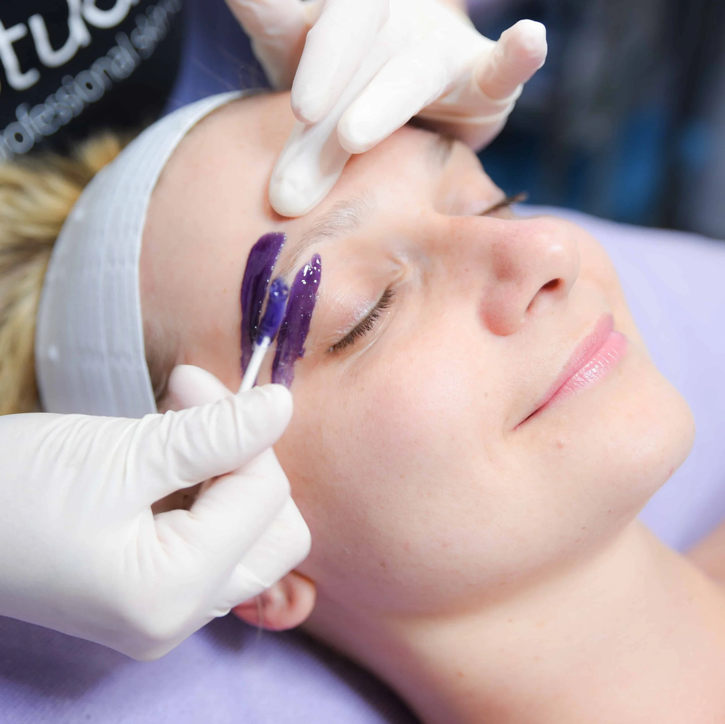 A woman receiving a cosmetic procedure on her eyebrows with purple dye applied, lying peacefully with her eyes closed, while a professional in gloves administers the treatment.