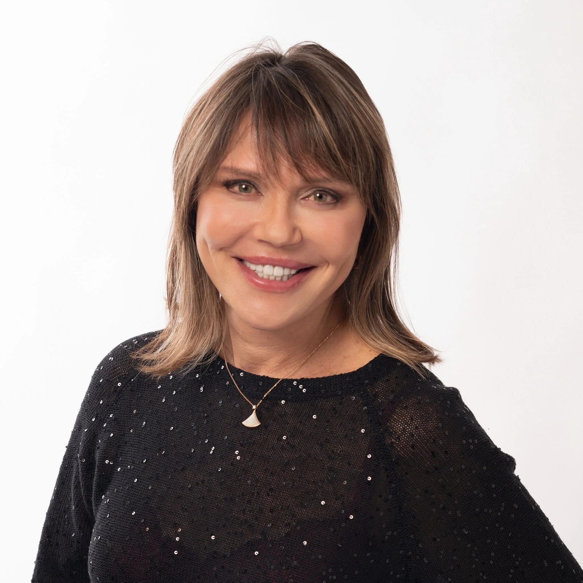 A woman with shoulder-length light brown hair wearing a black sequined top and a necklace with a fan-shaped pendant, smiling confidently against a white background.