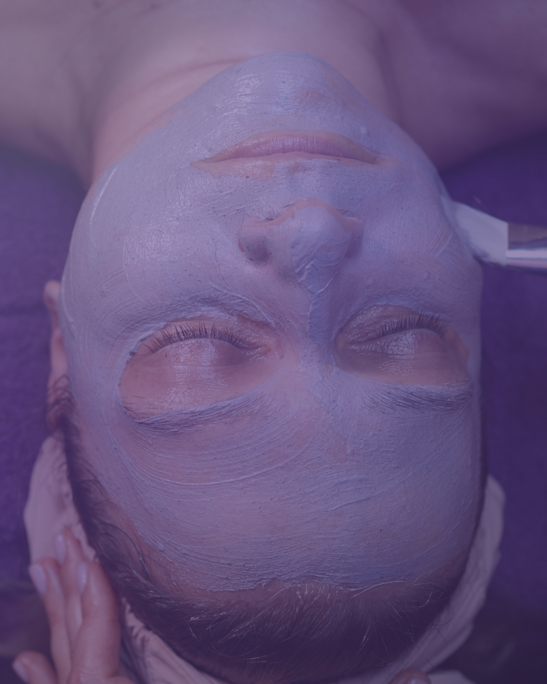 A person lying down with eyes closed while receiving a facial mask treatment, with a soft purple light over the scene.