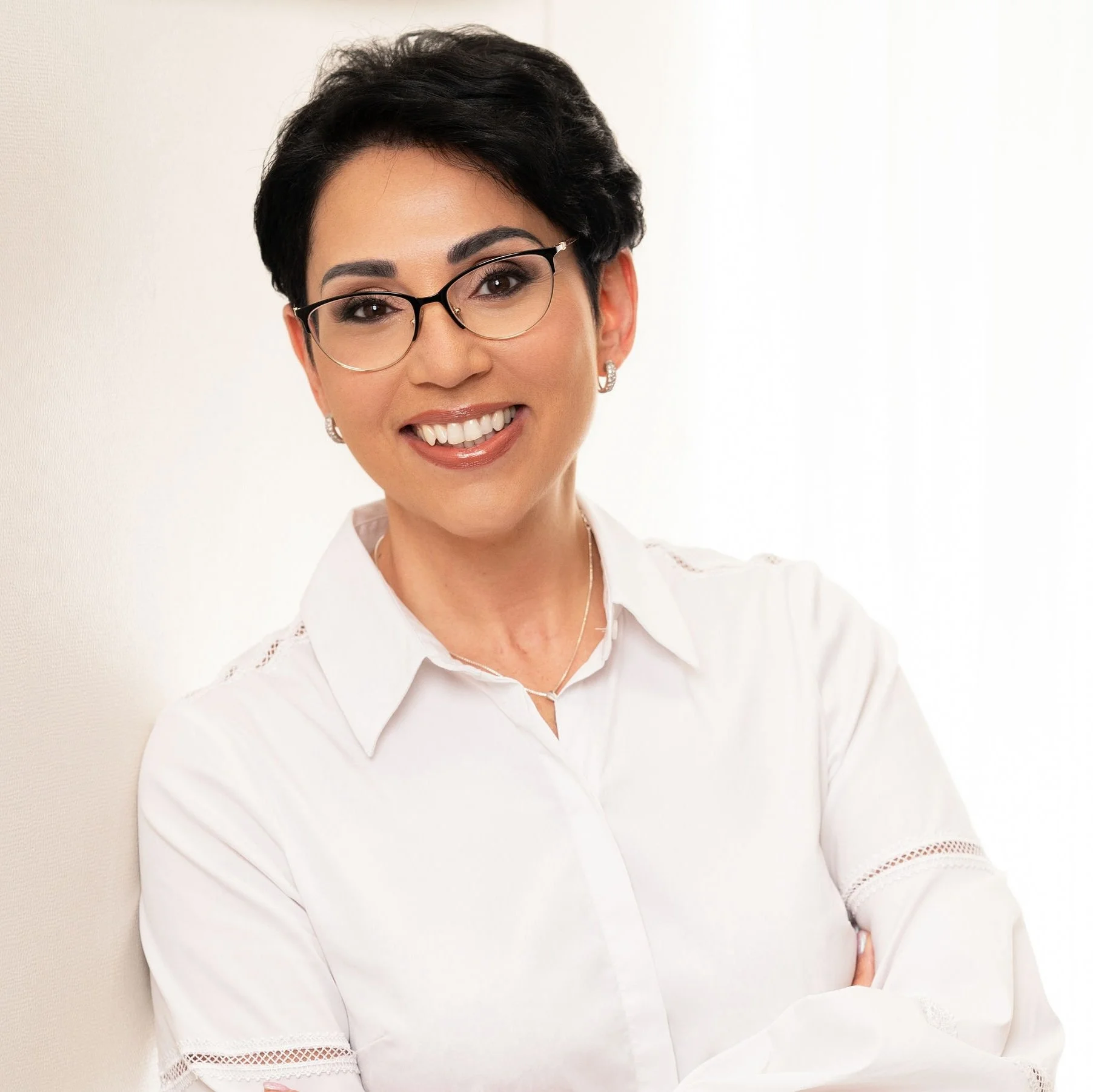 Smiling woman with short dark hair and glasses, wearing a white blouse, standing against a light background.