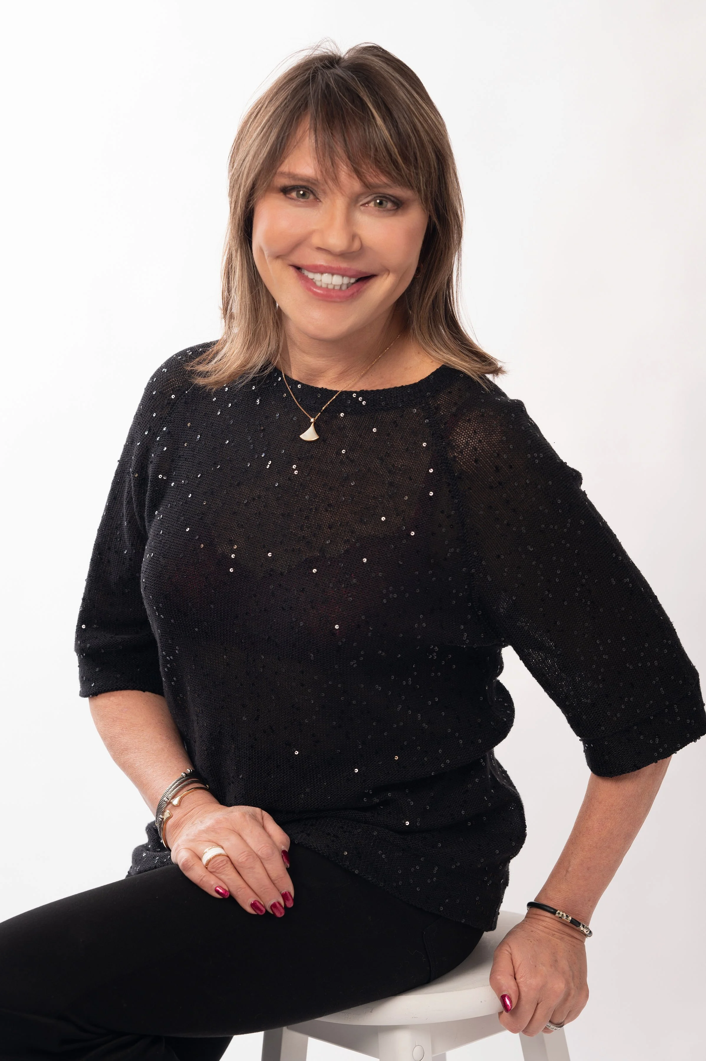 A woman with shoulder-length light brown hair, wearing a black, sparkly top with three-quarter sleeves, sitting on a white stool against a plain white background, smiling at the camera.