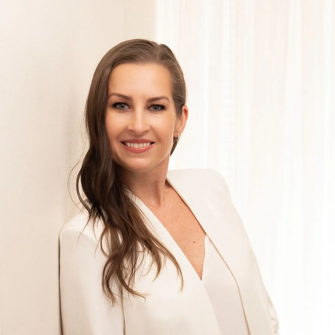 A woman with long, wavy brown hair smiling and leaning against a white wall, wearing a white blazer.