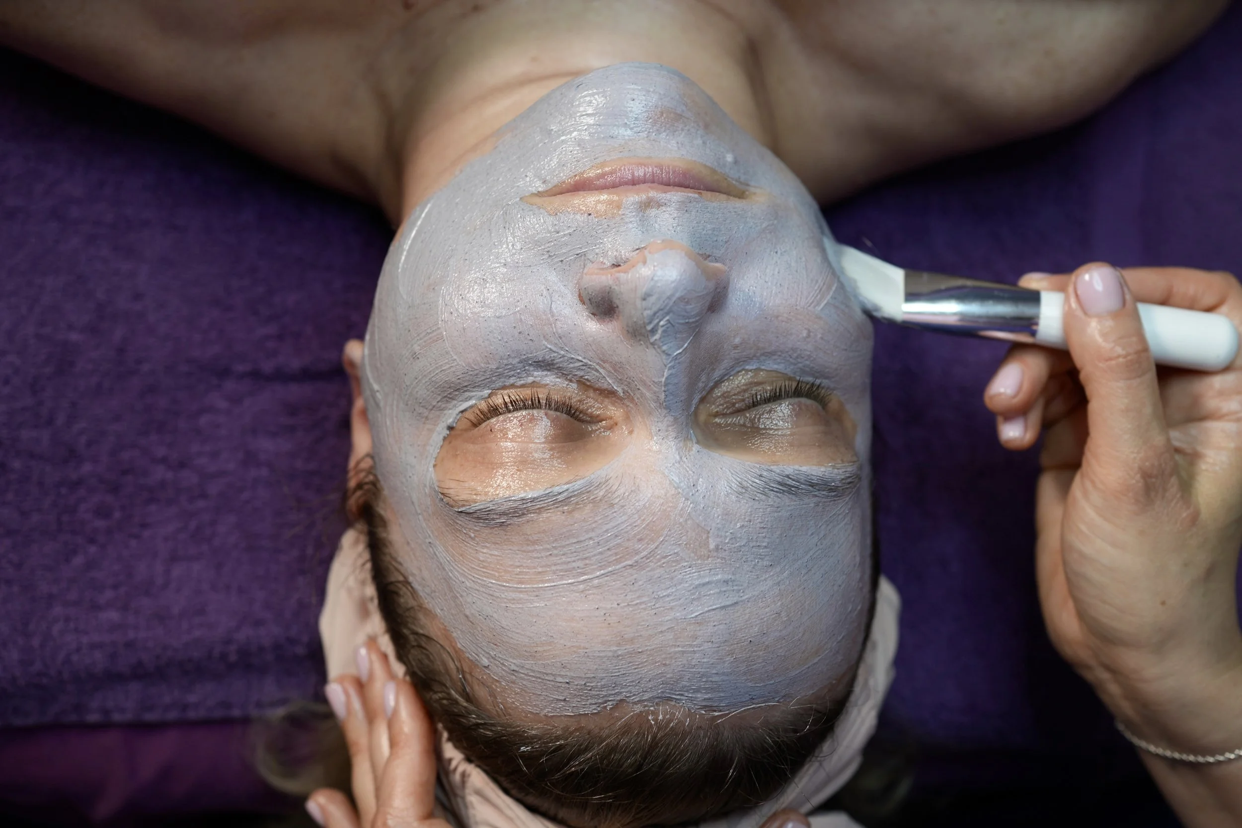 A woman is getting a facial treatment with a clay mask applied to her face while lying down.
