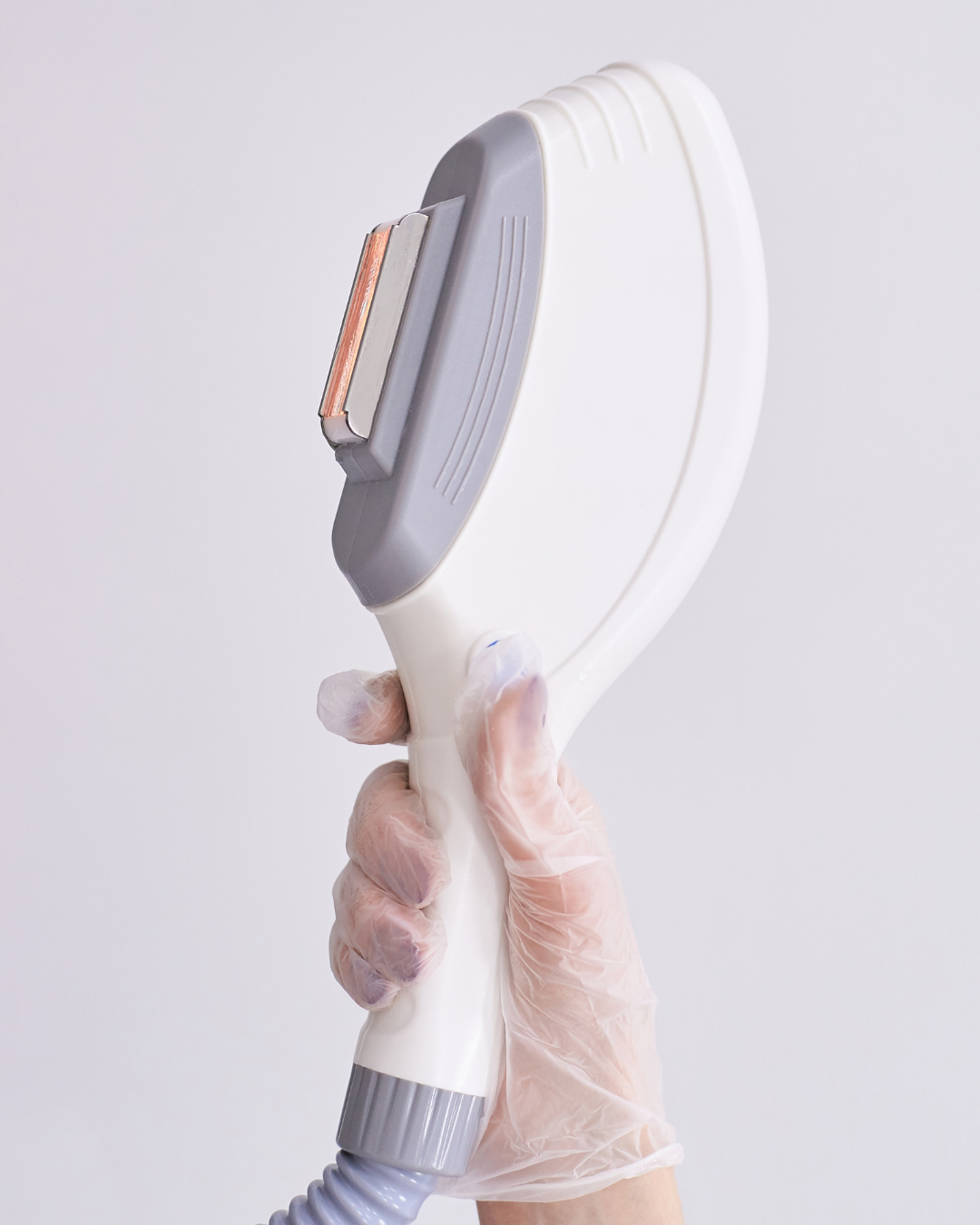 A hand wearing a transparent glove holding a white electronic device for IPL hair removal with a gray handle and a metallic blade head against a plain white background.