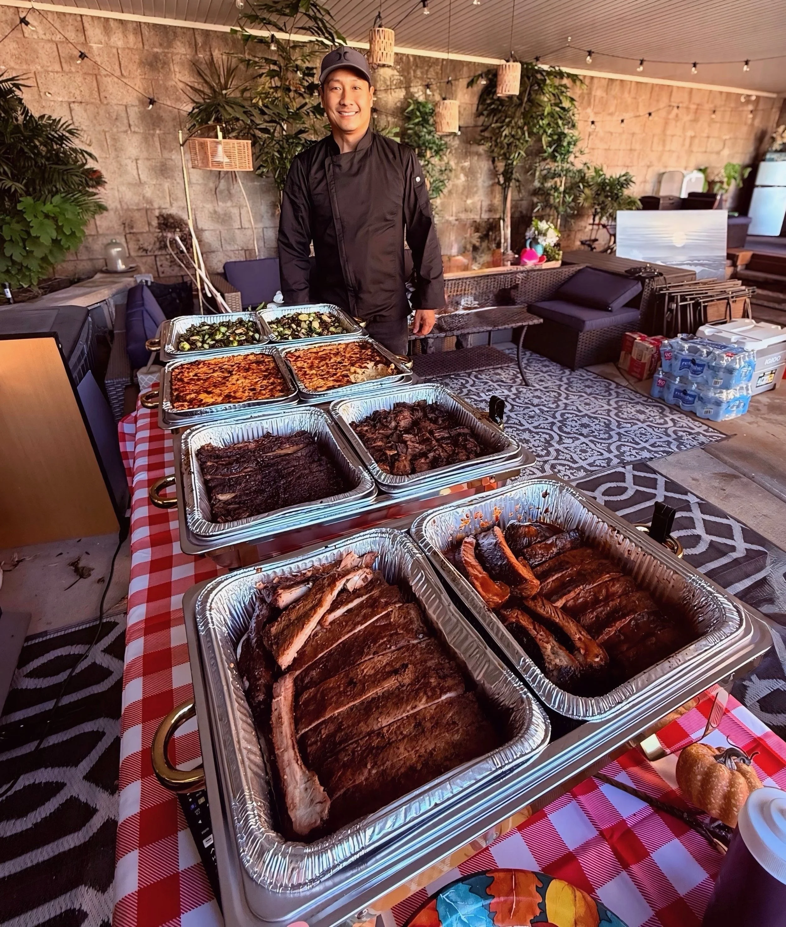 A man in a chef's jacket and cap standing behind a table with trays of barbecued meats and side dishes, set in a cozy outdoor patio area with plants, hanging lights, and patterned flooring.