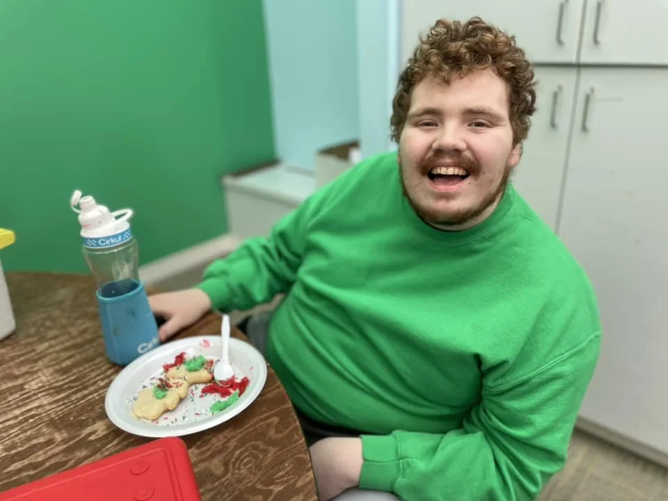 A young man with curly hair and a beard, wearing a bright green shirt, sitting at a wooden table with a plate of decorated sugar cookies, a reusable water bottle, and a red lunch container. He is smiling and in a kitchen with light-colored cabinets.