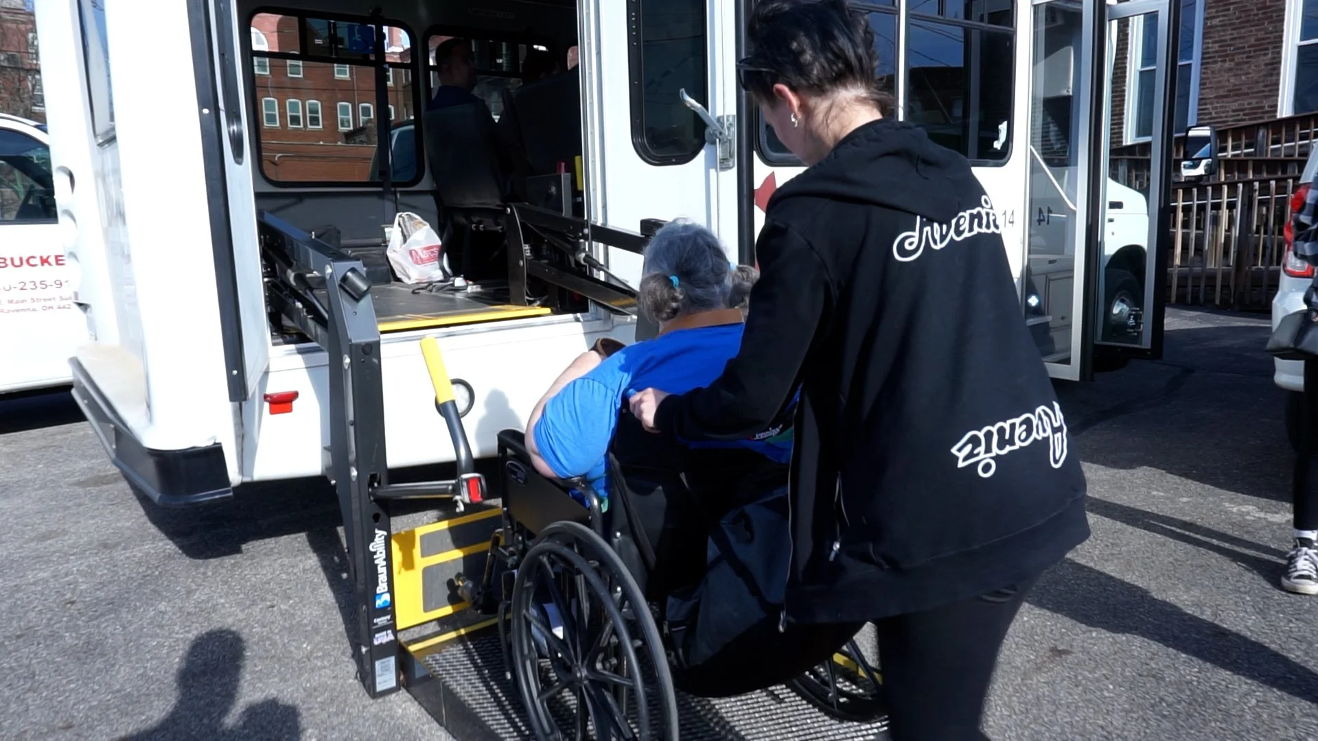 A person in a wheelchair being helped onto a food truck by a woman wearing a black hoodie with the word 'Duenia' on the back. The food truck has an accessibility lift, and there are other vehicles and buildings in the background.