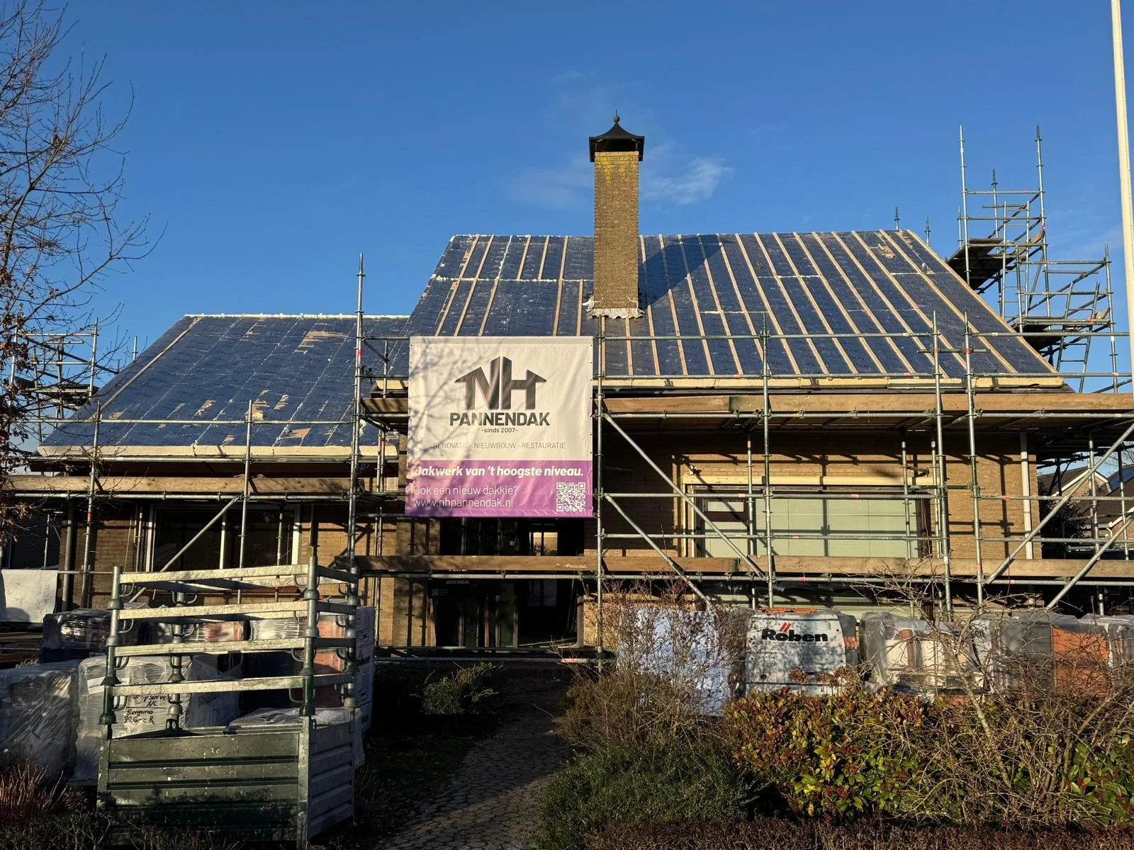 Construction of a house with exposed roof framing and scaffolding surrounding the building, with a sign for PannenDak company.