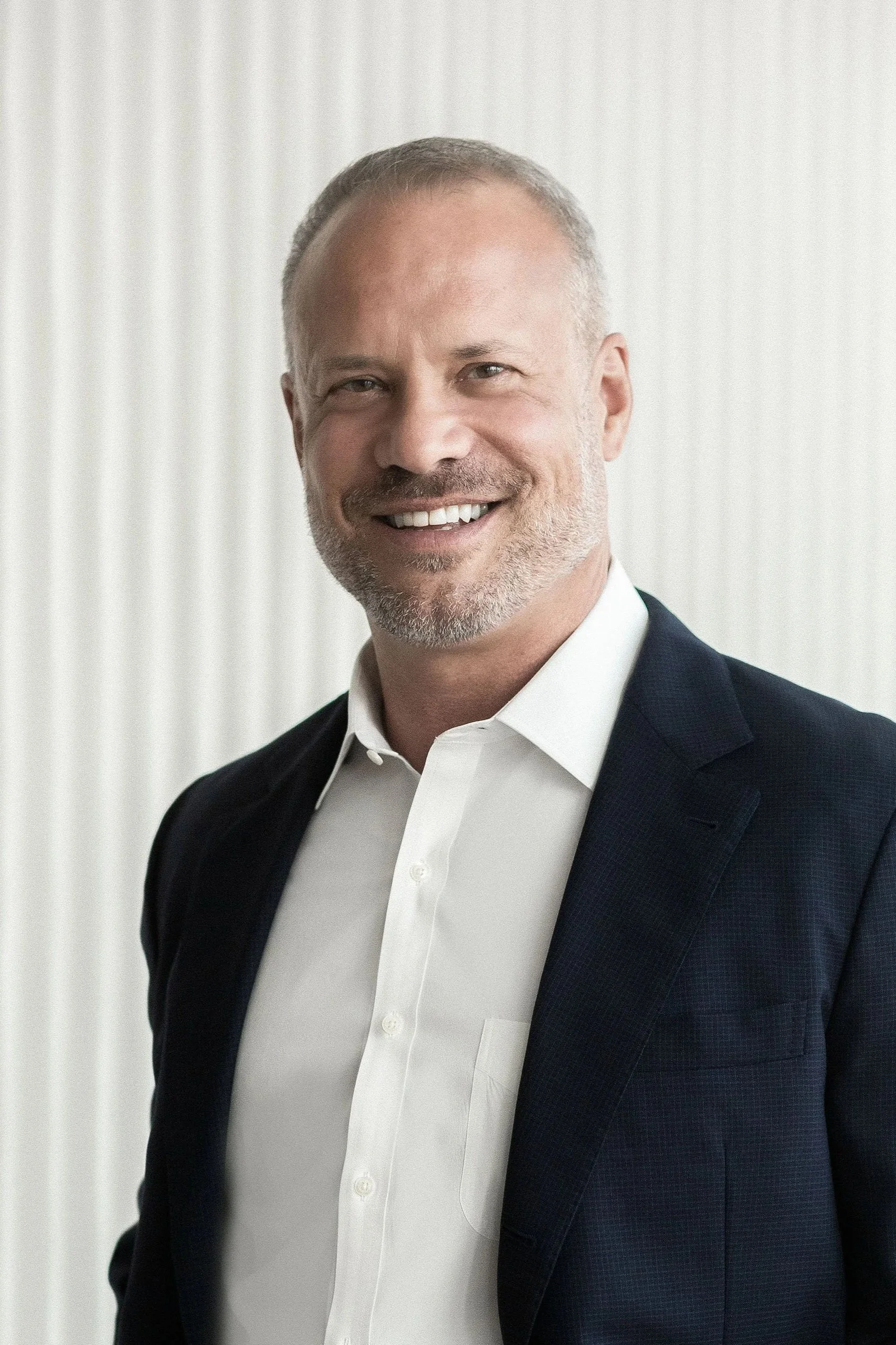 A portrait of man with gray hair and a beard, smiling, wearing a white dress shirt and a dark suit jacket, standing in front of a light-colored background.