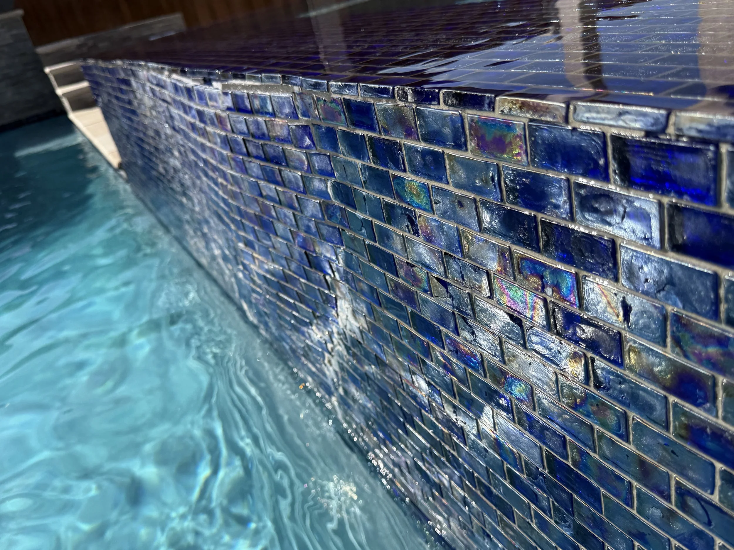Polished blue mosaic pool tile at waterline with sunlight reflections.
