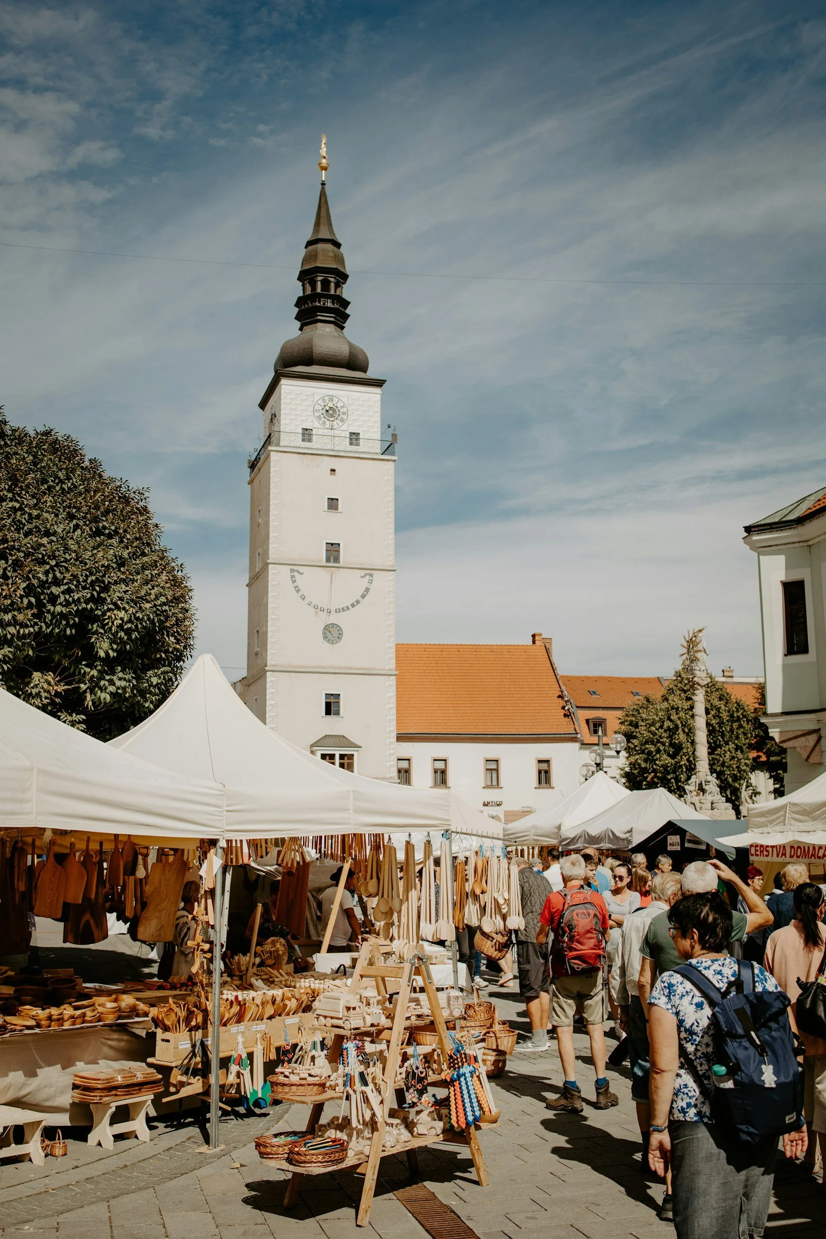 Outdoor market with tents selling wooden crafts, with a historic white clock tower and a crowd of people walking around.