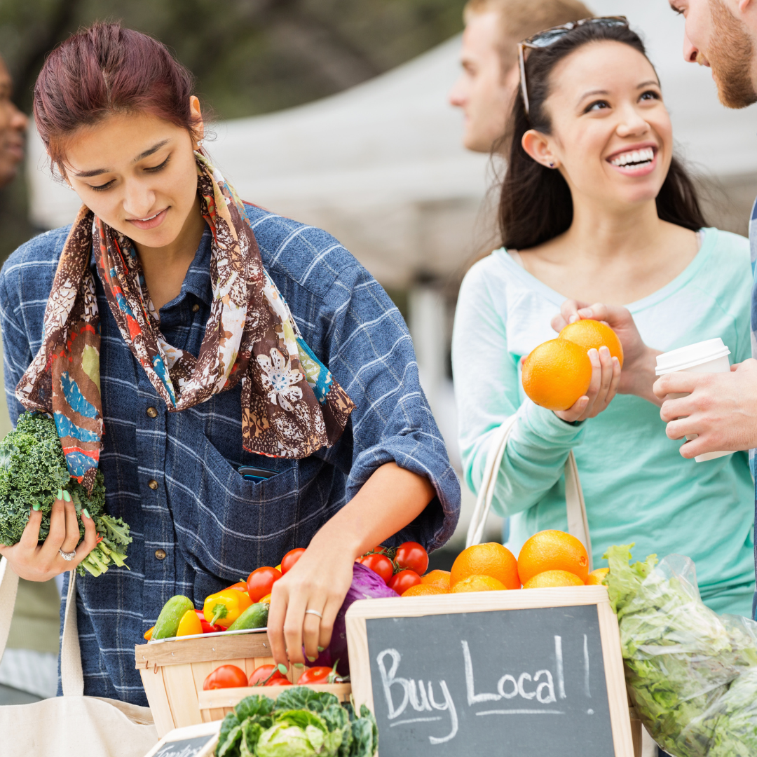 People at a farmer's market, with a woman selecting fresh vegetables and a sign that says 'Buy Local'.