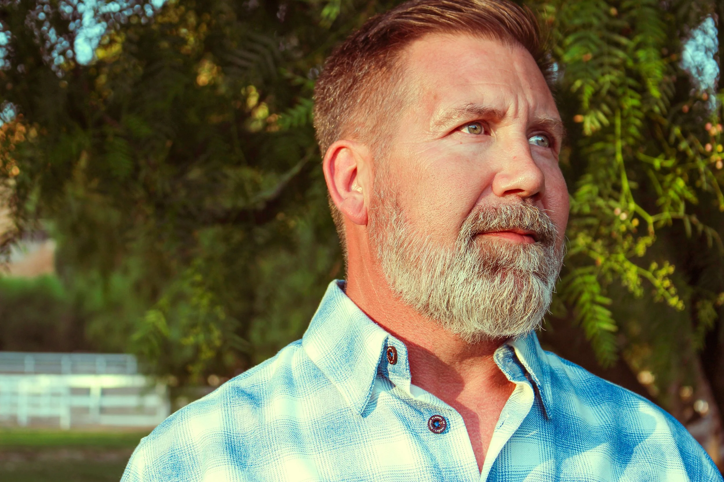 A middle-aged man with a beard and short brown hair standing outdoors in front of green foliage, looking off into the distance.