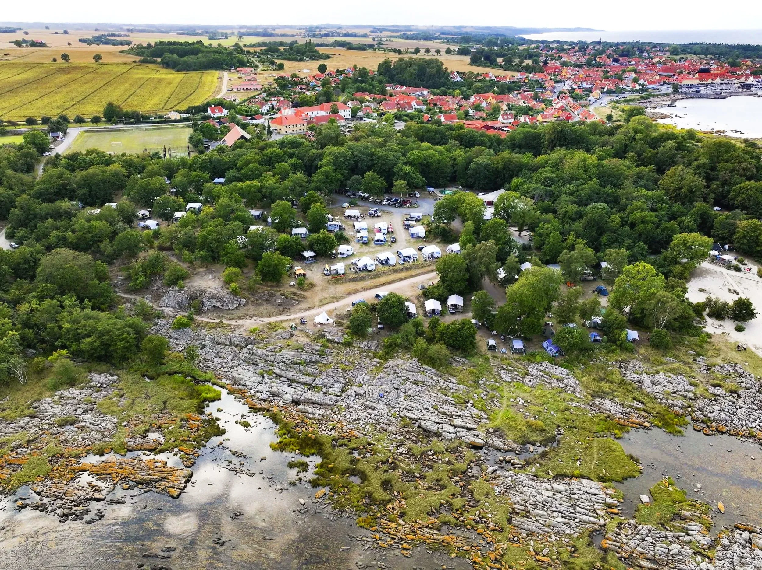 Luftfoto af en kystlinie med vand, sten, grøn vegetation, campingvogne, skov og en by med røde tage.