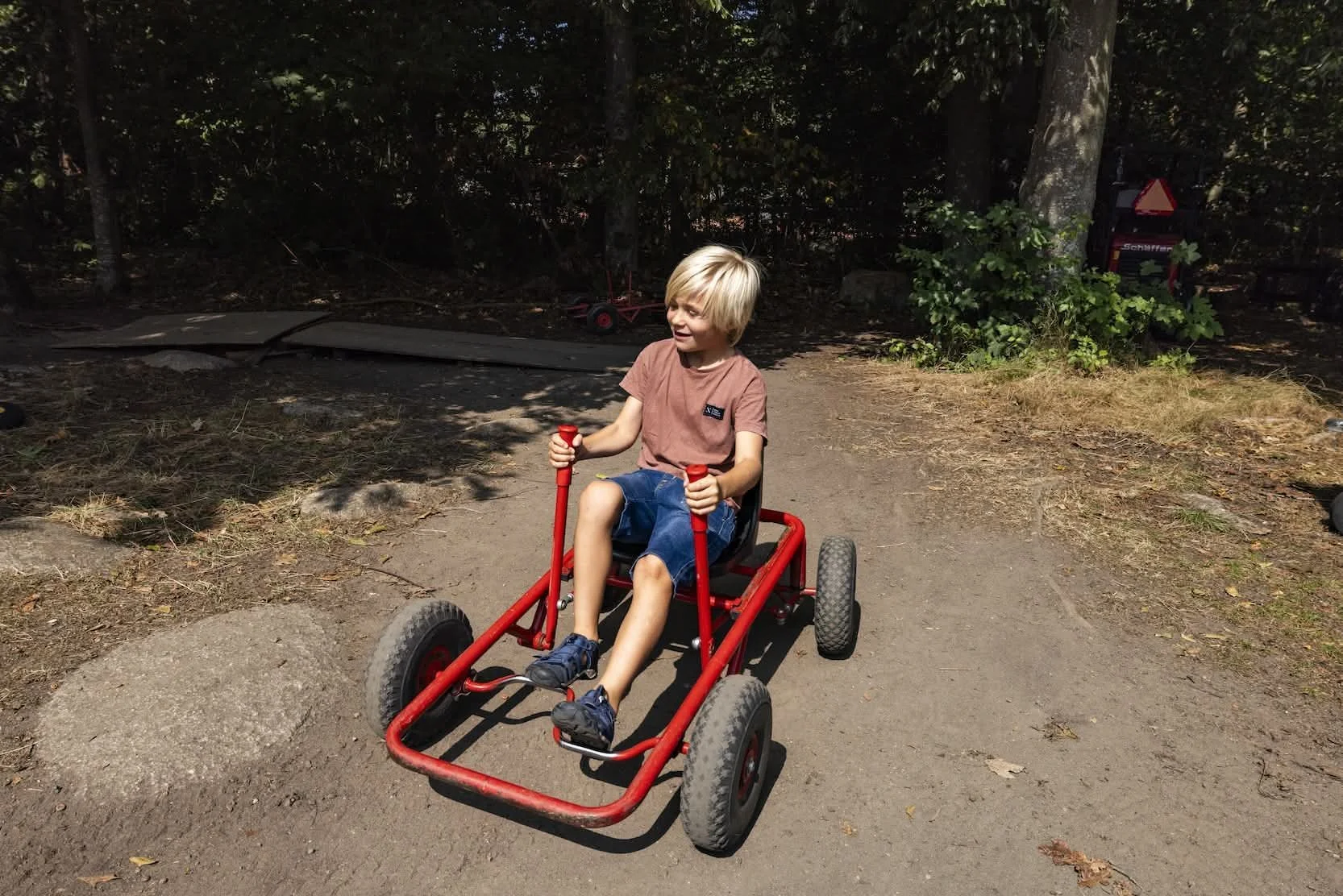 En dreng sidder i en rød bugatbil på en natursti, omgivet af træer og skygge.