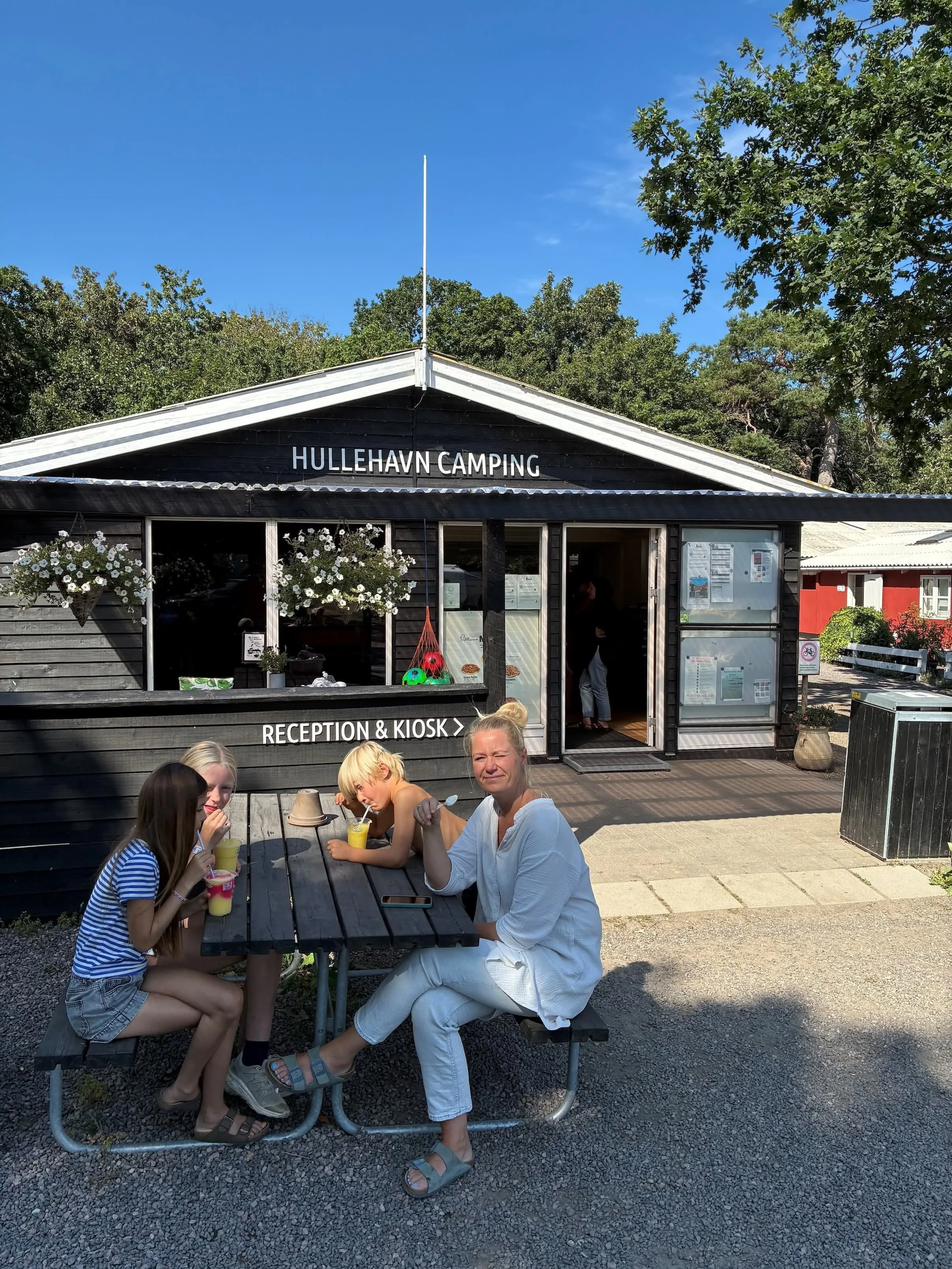 Familie sidder ved picnicbord foran receptionen på HulletHavn Camping, nyder drinks i solen.