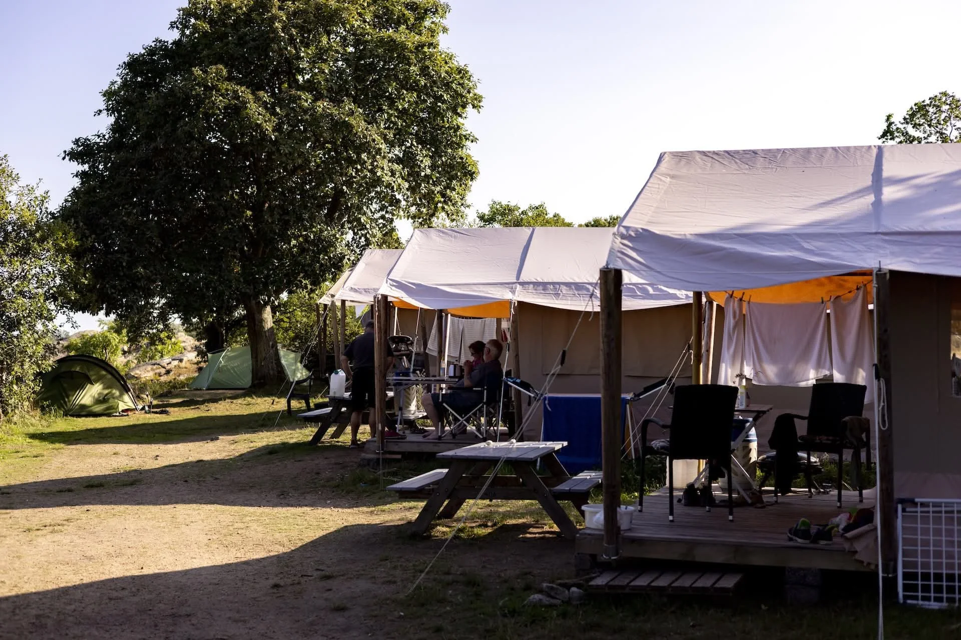 Campsite with tents and a wooden deck under a large tree, with camping gear and people relaxing.