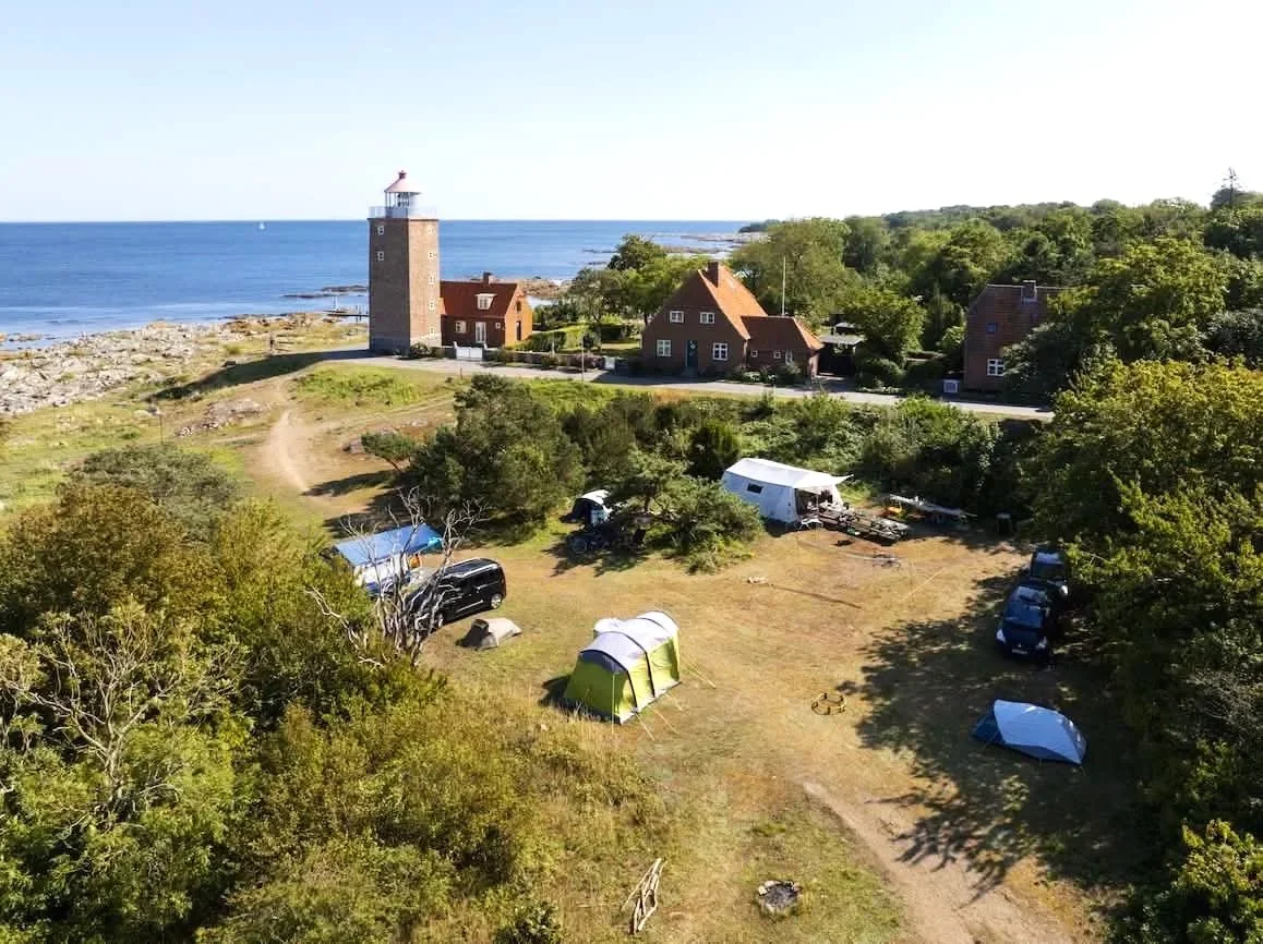 Strandnær campingplads med telte, biler og campervans, med en kystlinje med klipper og en fyrtårn i baggrunden, omgivet af grønne træer og en vej.