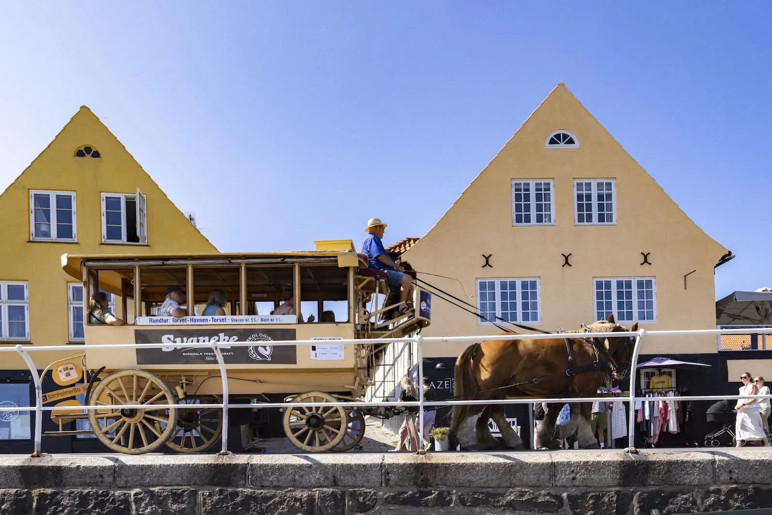 En hestevogn med passagerer kører langs en fortove i en by med gule bygninger under en klar blå himmel.
