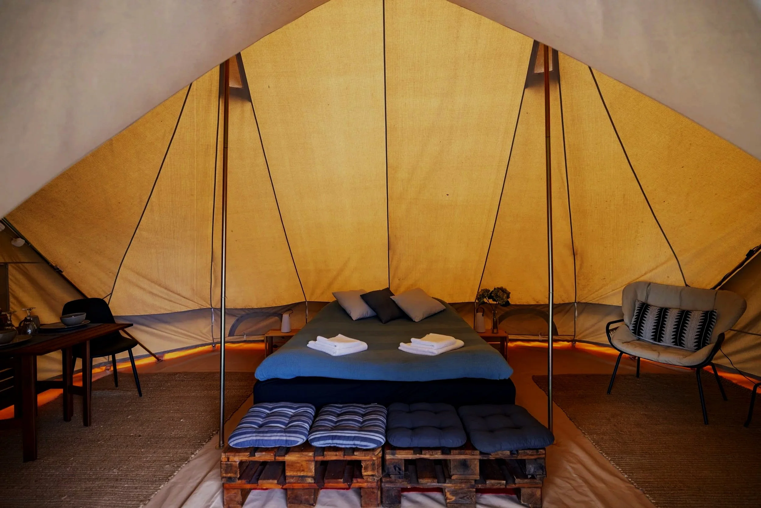 Indersiden af en glampingtelt med seng, pude og tæpper. Der er også et bord med service og en stol.
